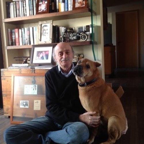 L'image montre un homme assis sur le sol, avec un chien à ses côtés. L'homme porte un pull noir et a des cheveux grisonnants. Le chien, de taille moyenne, a un pelage brun et semble calme. En arrière-plan, on peut voir une bibliothèque remplie de livres, ainsi que quelques objets personnels comme des cadres photo et des souvenirs. L'ambiance de la pièce paraît chaleureuse et accueillante.