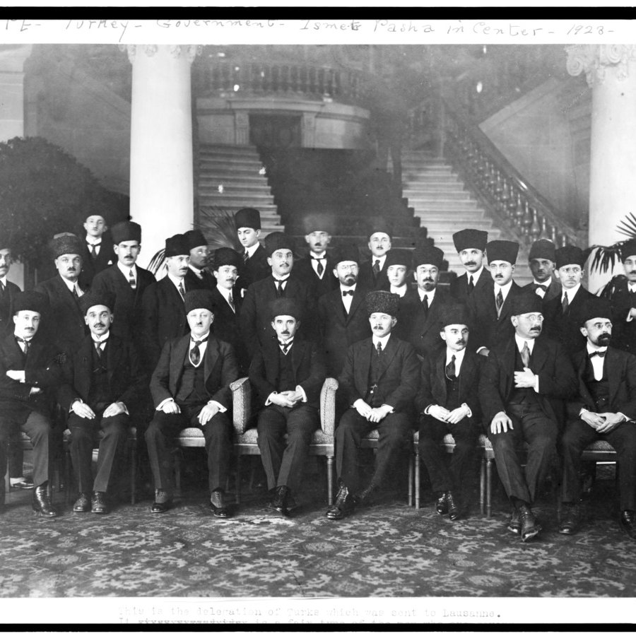 L'image présente un groupe d'hommes, probablement d'importance politique ou officielle, assis et debout dans un cadre formel. Ils portent des costumes sombres et des chapeaux traditionnels, et certains ont des moustaches. La scène se déroule dans un intérieur élégant, avec des colonnes décoratives et des plantes en pot en arrière-plan. L'atmosphère dégage une impression de solennité et de cérémonie, suggérant un événement ou une réunion importante.