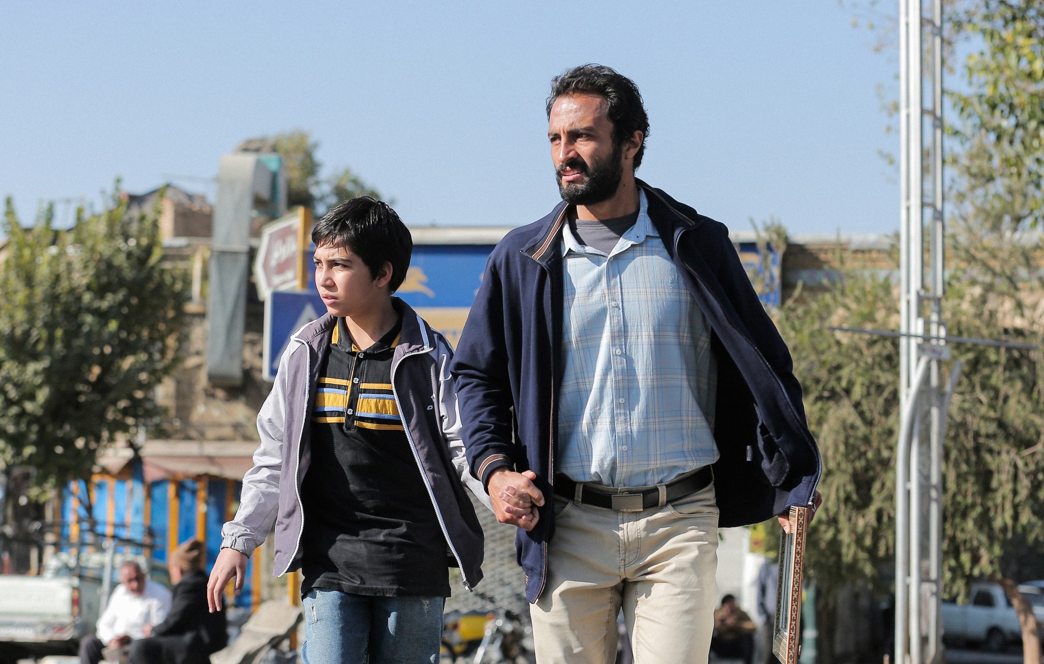 L'image montre un homme et un garçon marchant ensemble dans une rue. L'homme, qui semble être un adulte, tient la main du garçon, qui est jeune et porte un haut à rayures. En arrière-plan, on aperçoit des bâtiments et des arbres, ce qui suggère un environnement urbain. L'éclairage est clair, indiquant probablement une belle journée. L'atmosphère semble à la fois tendre et protectrice, mettant en avant la relation entre les deux personnages.