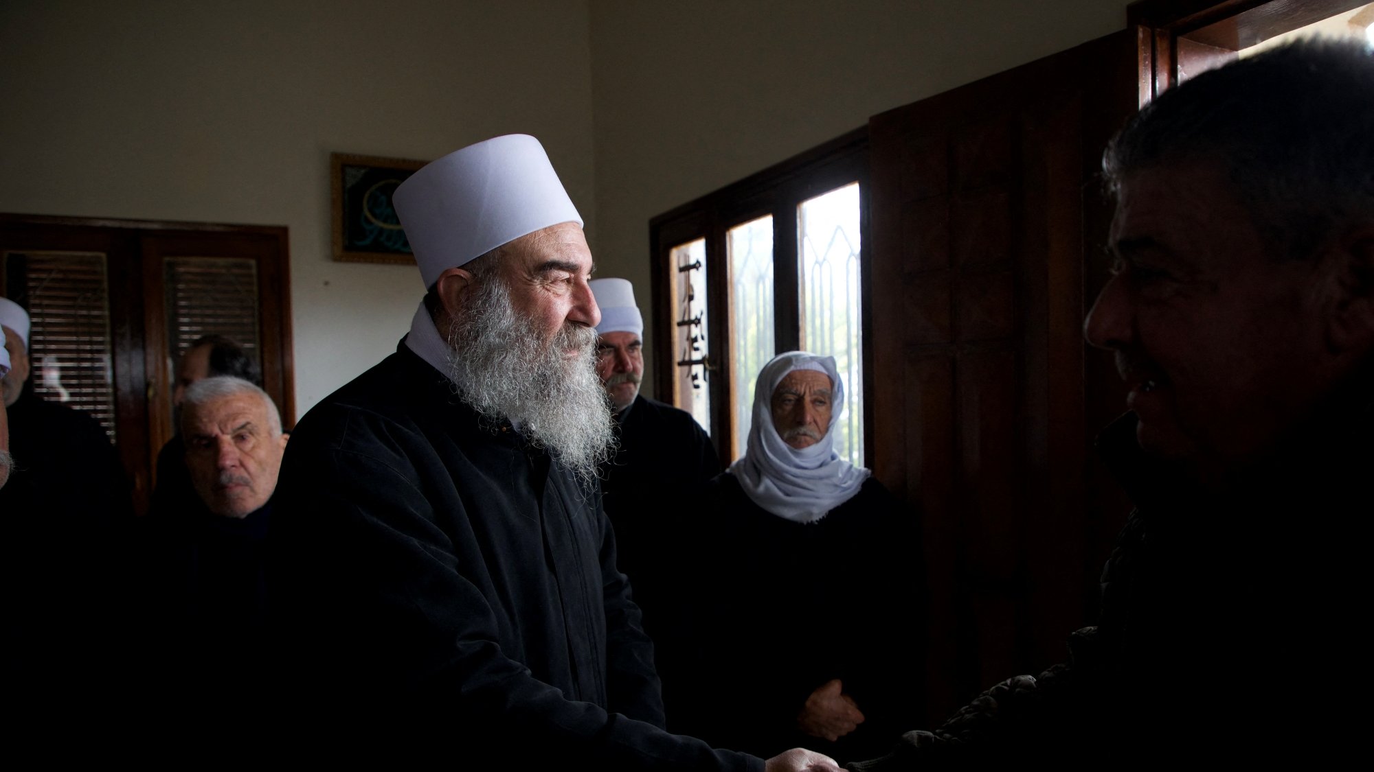 Group of men, some with white hats and beards, shaking hands and wearing dark clothing in a room with windows.