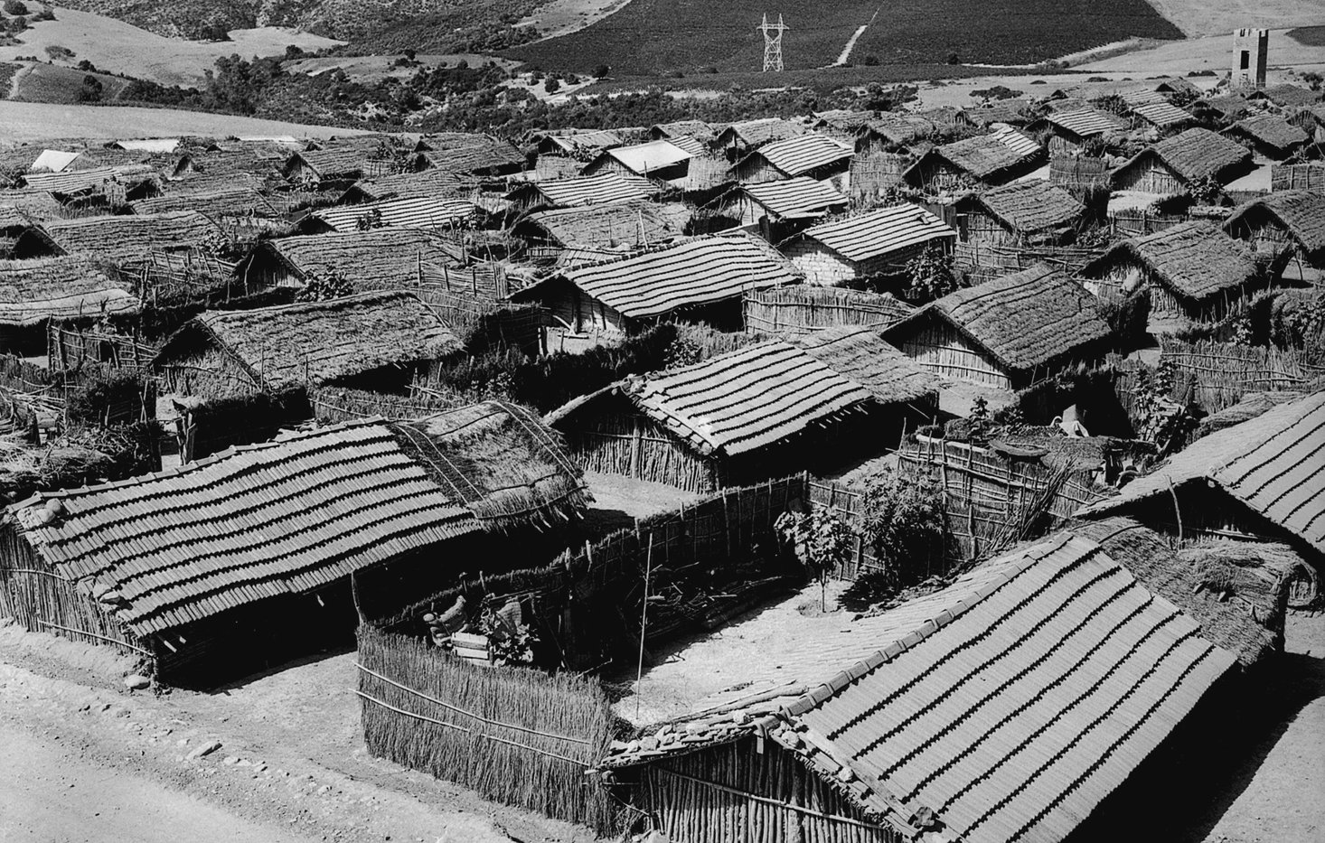 L'image montre un paysage rural avec plusieurs maisons en bois et en matériau naturel, probablement des huttes, disposées de manière dense. Les toits sont recouverts de paille ou d'un matériau similaire. On aperçoit également des collines verdoyantes à l'arrière-plan, ce qui suggère un environnement rural. Le ciel est clair, et l'ensemble de la scène a une tonalité noire et blanche, conférant une atmosphère nostalgique et ancienne à l'image.