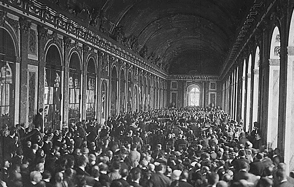 L'image montre une vaste salle intérieure, probablement un palais, remplie d'un grand nombre de personnes. On peut voir des rangées de chaises disposées, et les murs sont ornés de grandes fenêtres et de détails architecturaux. La foule semble rassemblée pour un événement ou une cérémonie importante, créant une atmosphère dense et dynamique dans cet espace majestueux. La lumière pénètre par les fenêtres, ajoutant une ambiance solennelle à la scène.