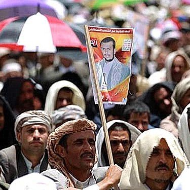 L'image montre une grande foule de personnes réunies, probablement lors d'une manifestation ou d'un rassemblement. La majorité des participants portent des vêtements traditionnels et certains sont abrités sous des parapluies de différentes couleurs. Au premier plan, un homme tient une affiche avec un portrait, ce qui suggère qu'il soutient une personnalité ou une cause spécifique. L'atmosphère semble engagée et collective.