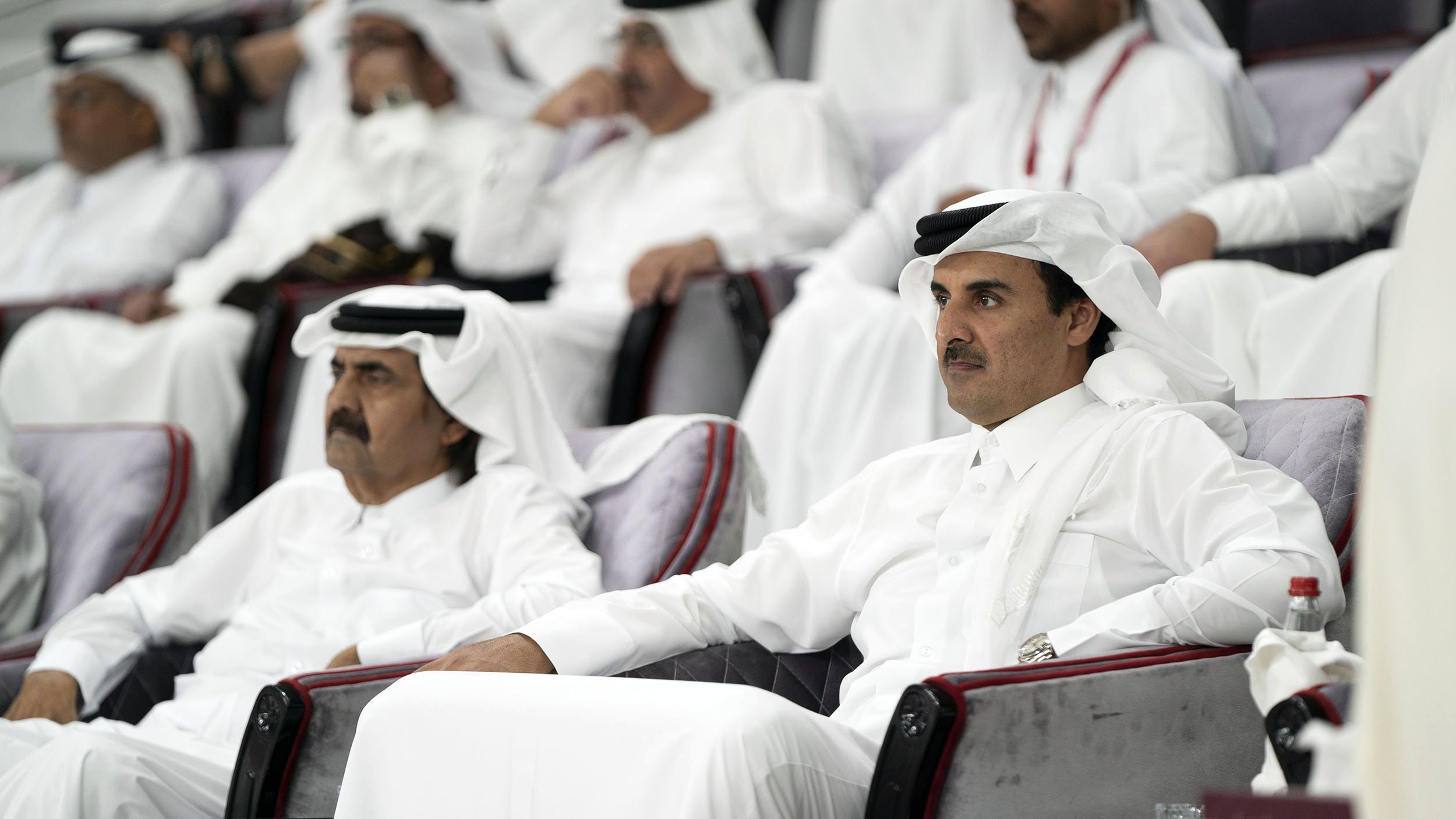 L'image montre un groupe de personnes assises dans des sièges, probablement dans un stade ou un lieu événementiel. Deux hommes sont au premier plan, vêtus de vêtements traditionnels qataris, avec des turbans blancs et des robes longues. Ils semblent attentifs et engagés dans l'événement se déroulant devant eux. L'atmosphère est celle d'un rassemblement formel, avec d'autres personnes en arrière-plan également habillées de manière traditionnelle.