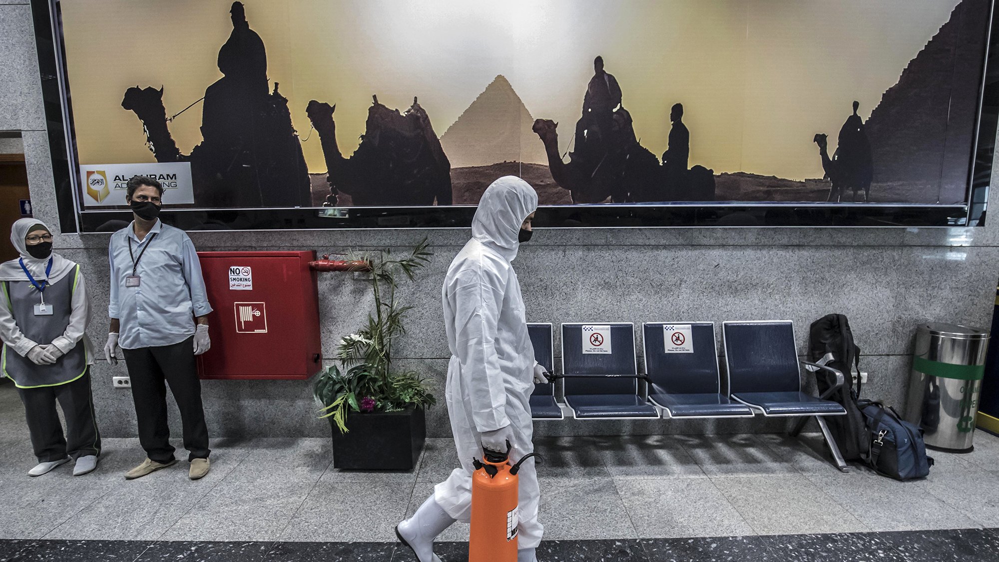 The image depicts a scene in a public area, likely an airport or a similar facility. In the foreground, a person wearing a white protective suit and mask is walking while holding an orange container. In the background, a large advertisement features silhouettes of camels and a pyramid, suggesting a connection to Egypt. There are two individuals in the background, also wearing masks, standing near a fire extinguisher. The seating area appears empty, adding to a sense of quietness in the environment.
