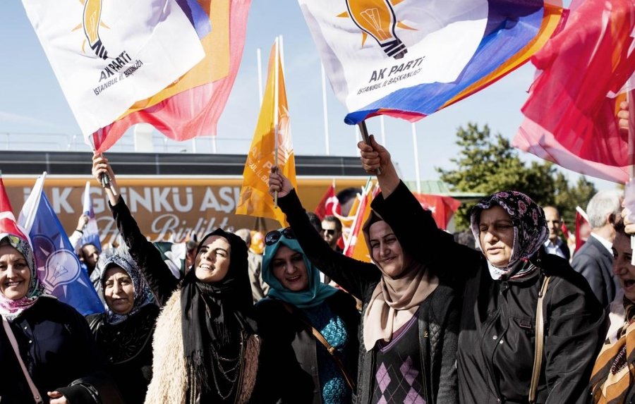 L'image montre un groupe de femmes rassemblées lors d'un événement ou d'une manifestation, brandissant des drapeaux. Ces drapeaux portent le logo d'un parti politique, probablement le Parti de la justice et du développement (AK Parti) en Turquie. Les femmes semblent enthousiastes et engagées, portant des vêtements variés, y compris des foulards. L'arrière-plan présente d'autres drapeaux et des affiches, créant une ambiance festive et militante.