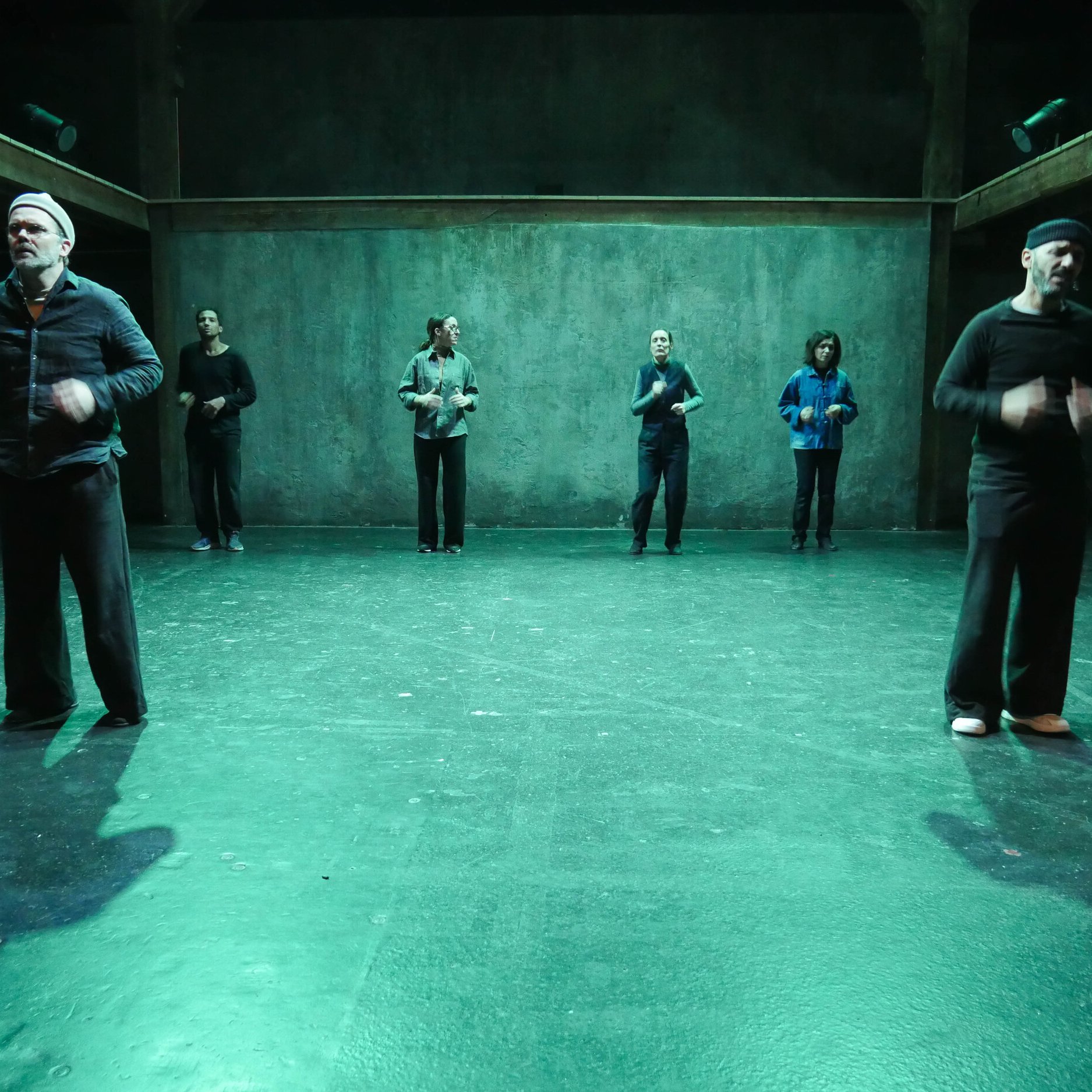 L'image montre un groupe de six personnes sur scène, dans un espace artistique éclairé d'une lumière verte. Elles semblent engagées dans une performance ou une répétition. Les membres du groupe portent des vêtements de différents styles, et se déplacent à divers endroits de la scène. L'arrière-plan est dépouillé, avec des murs en béton, ce qui crée une ambiance minimaliste et contemporaine.