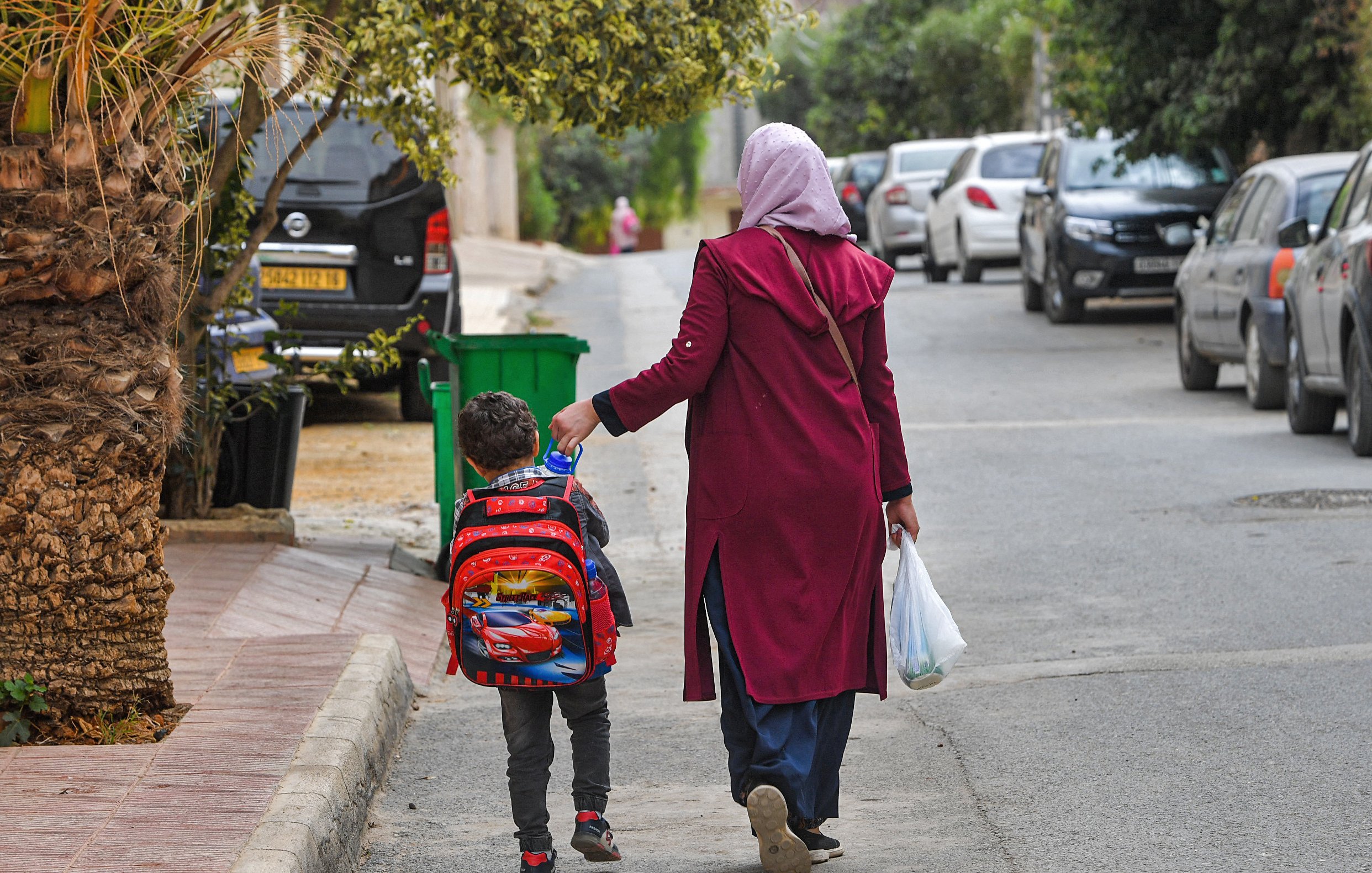L'image montre une femme portant un foulard, vêtue d'un long manteau, qui marche dans une rue résidentielle avec un jeune garçon. Le garçon porte un sac à dos coloré, probablement inspiré d'un personnage de dessin animé. Ils semblent se diriger vers une destination, et la femme tient également un sac en plastique. En arrière-plan, on peut voir des voitures garées des deux côtés de la rue et de la verdure autour. L'atmosphère semble calme et quotidienne.