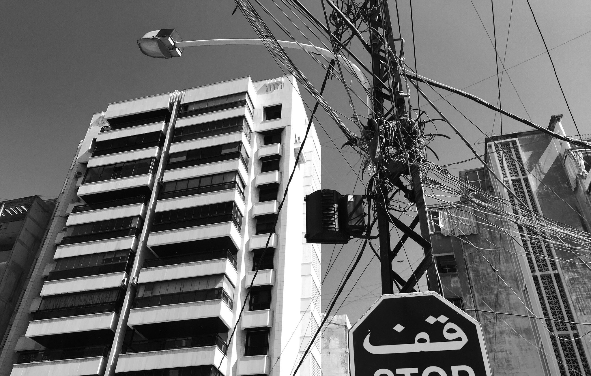 این تصویر یک خیابان شهری را نشان می‌دهد که در آن یک تابلو ایست با نوشته "قف" (STOP) قرار دارد. در پس‌زمینه، یک ساختمان بلند و مدرن به همراه خطوط برق در آسمان دیده می‌شود. رنگ تصویر سیاه و سفید است که به آن حال و هوای خاصی می‌بخشد. ظاهر شهر و معماری ساختمان‌ها، نمایی از زندگی روزمره شهری را ارائه می‌دهد.