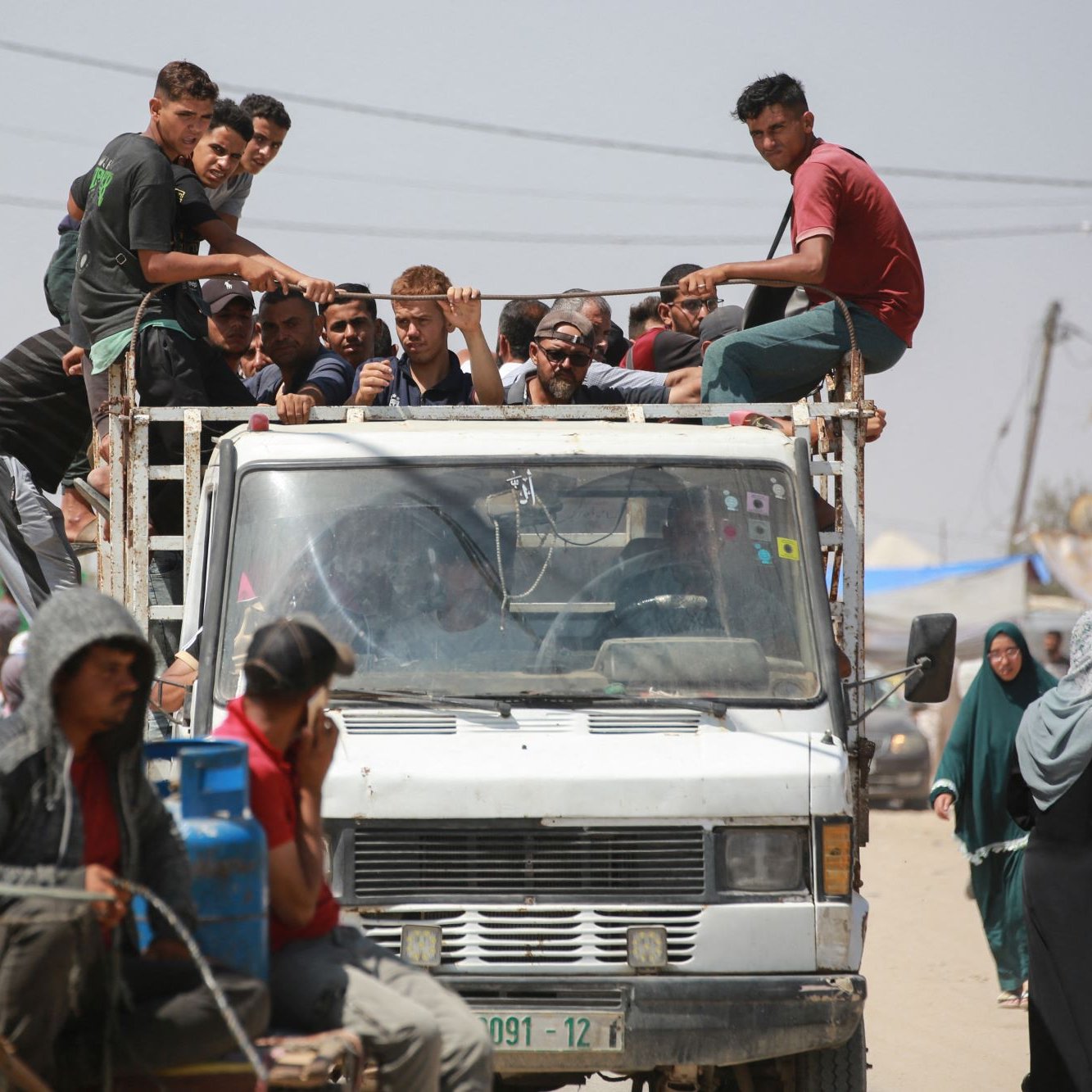 در این تصویر، گروهی از جوانان در حال نشستن بر روی یک کامیون هستند. برخی از آن‌ها در قسمت بار کامیون نشسته‌اند، در حالی که دیگران به طرف خودرویی که در حرکت است، نگاه می‌کنند. در جلوی تصویر، چند نفر در کنار کامیون و همچنین زنانی با پوشش‌های سنتی مشاهده می‌شوند. در پس‌زمینه، سایه‌هایی از چادرها و همچنین سیم‌کشی‌های برق دیده می‌شود که نشان‌دهنده یک منطقه پرجمعیت و شاید غیررسمی است. احساسی از شلوغی و جنب و جوش از صحنه به‌طور کلی مشهود است.