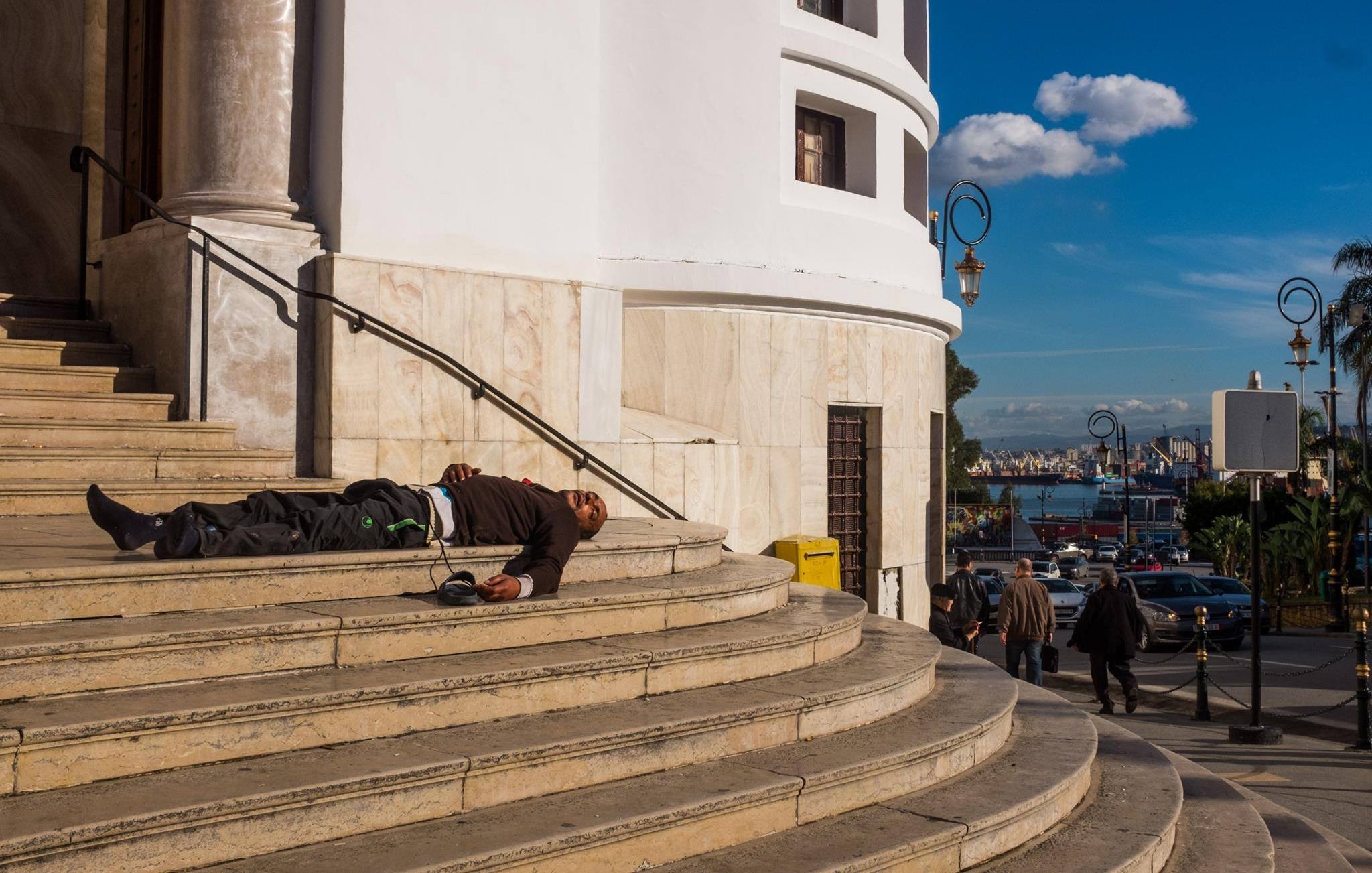 L'image montre un homme allongé sur les marches d'un bâtiment au style architectural classique, probablement en train de dormir ou en état de détresse. Le bâtiment est bien éclairé et ses murs sont de couleur claire, avec des colonnes visibles. En arrière-plan, on peut apercevoir une ville animée, avec des gens qui marchent et un port. Le ciel est bleu avec quelques nuages, ce qui donne une ambiance contrastée à la scène.