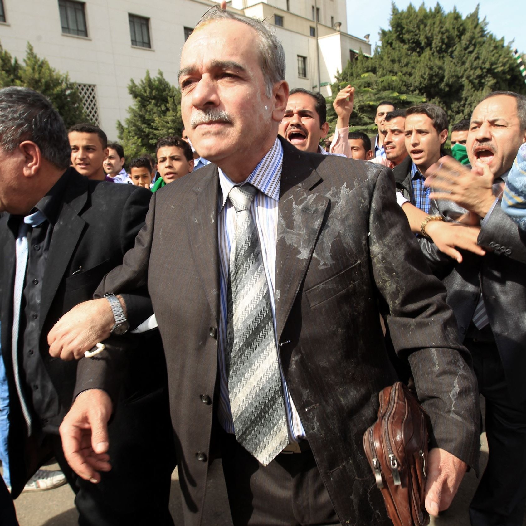 L'image montre un groupe de personnes dans une scène dynamique et animée. Au centre, un homme en costume, portant une cravate, semble marcher avec détermination, bien qu'il ait des éclaboussures sur son vêtement. Il est entouré de plusieurs autres individus qui semblent agités et expriment des émotions fortes, peut-être en criant ou en gesticulant. L'arrière-plan montre un environnement urbain avec des bâtiments et des arbres. L'atmosphère semble chargée, suggérant une situation de tension ou de protestation.