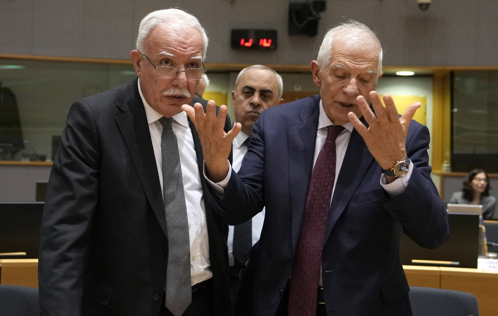 En la imagen se pueden ver a dos hombres en un entorno de reunión, posiblemente en una conferencia o un encuentro diplomático. Ambos parecen estar en una conversación intensa, con gestos de las manos que indican expresión o énfasis en lo que están discutiendo. Al fondo, hay otra persona observando. El ambiente es formal, con un espacio de trabajo y asientos visibles.