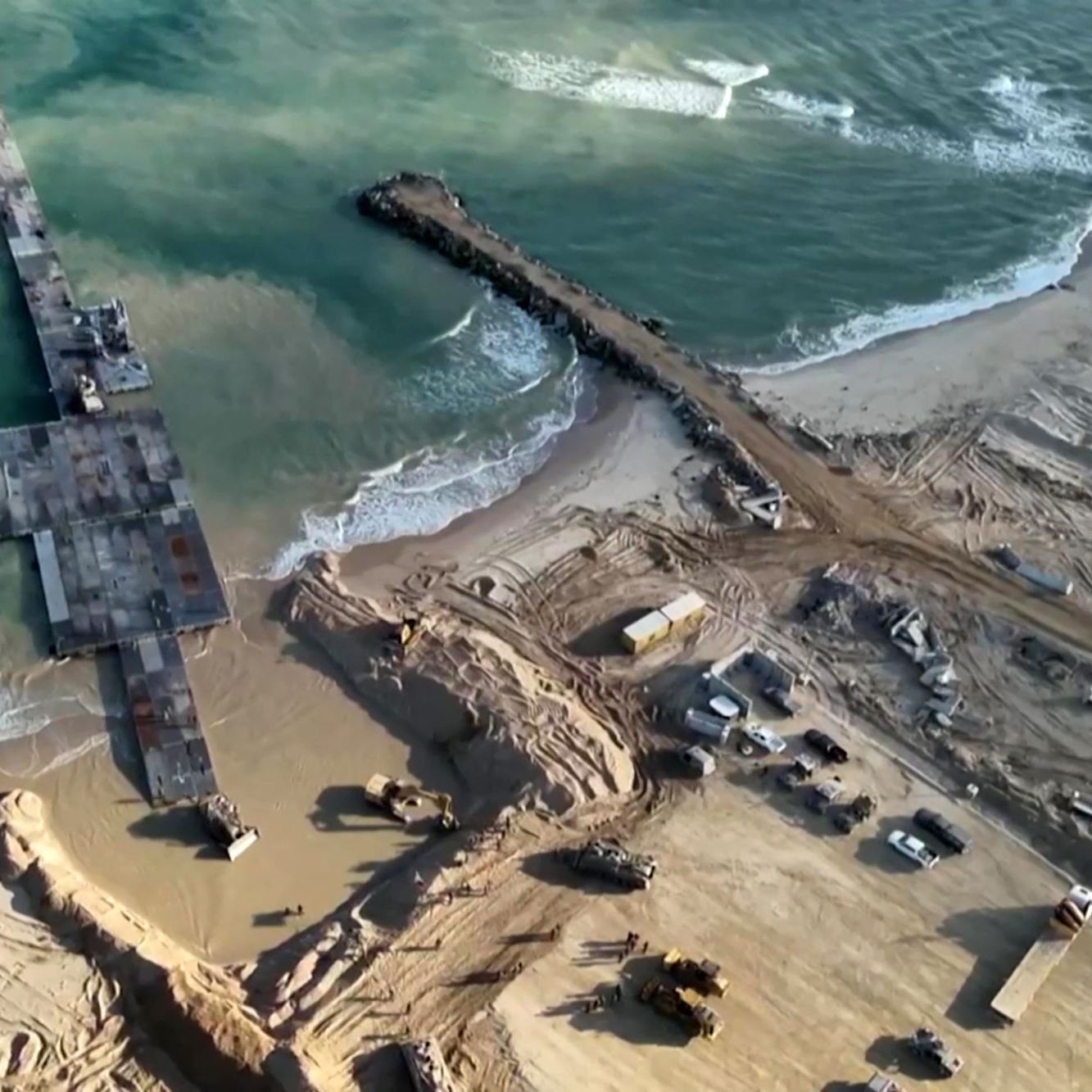 L'image montre une vue aérienne d'un site de construction situé près de la mer. On peut voir un port ou une jetée inachevée s'étendant dans l'eau, entouré de terres travaillées. Des engins de construction sont dispersés autour, et le sol est marqué par des travaux de terrassement. Les vagues de la mer s'échouent à proximité, créant un contraste avec la zone de construction. L'ensemble de la scène illustre une zone en développement, probablement pour une infrastructure côtière.
