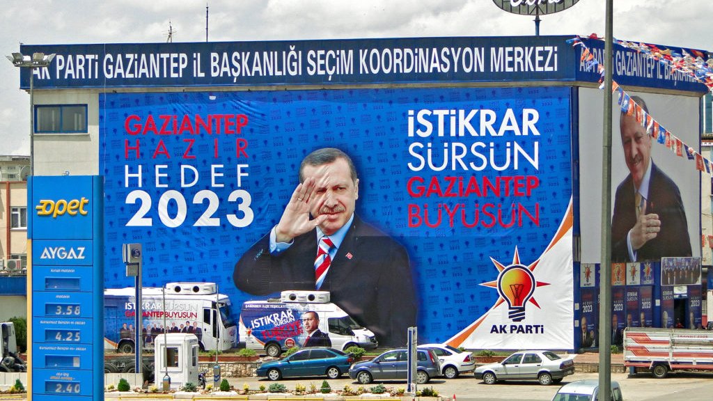 L'image montre un grand panneau publicitaire avec des slogans politiques. On y voit des phrases comme "GAZİANTEP HAZIR 2023" et "İSTİKRAR SÜRSÜN GAZİANTEP BÜYÜSÜN", suggérant une campagne électorale pour les élections de 2023. L'image présente également une figure politique qui salue, ajoutant une dimension dynamique à la publicité. Le fond inclut des symboles ou logos associés à un parti, probablement le parti AK. La scène est typique des campagnes électorales visuelles, avec une mise en avant forte du message et de la personnalité politique.