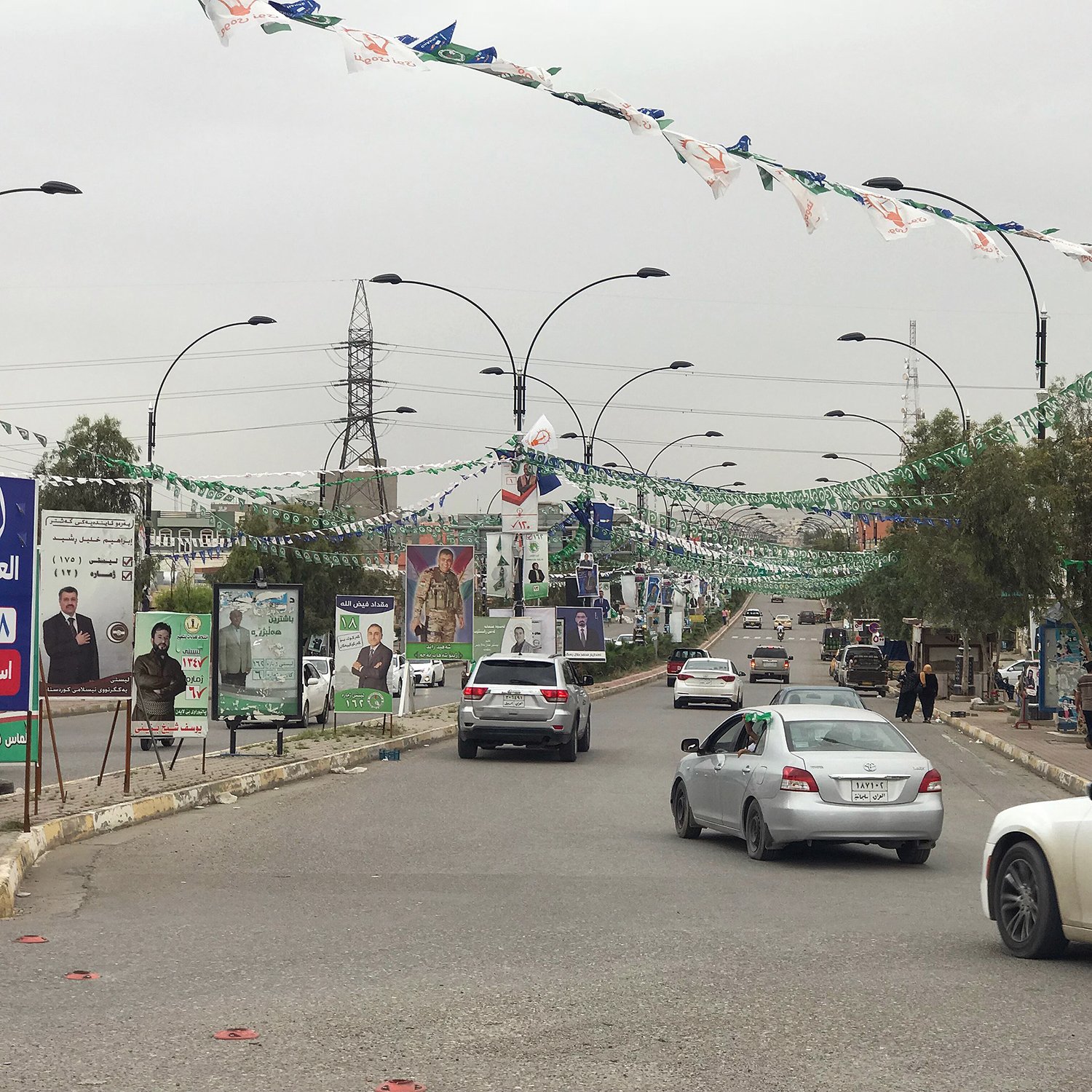 تظهر الصورة مشهداً لشارع في منطقة حضرية، حيث تنتشر الأعلام والزينة على أعمدة الإنارة. يمكن رؤية عدد من السيارات تسير على الطريق، مما يدل على حركة المرور. كما يوجد ملصقات وصور على الجدران، تشير إلى حملات انتخابية أو مناسبات خاصة. السماء تبدو ملبدة بالغيوم، مما يعطي انطباعاً عن جو غير مشمس. الأشجار والنباتات تضيف لمسة من الخضرة إلى المشهد.