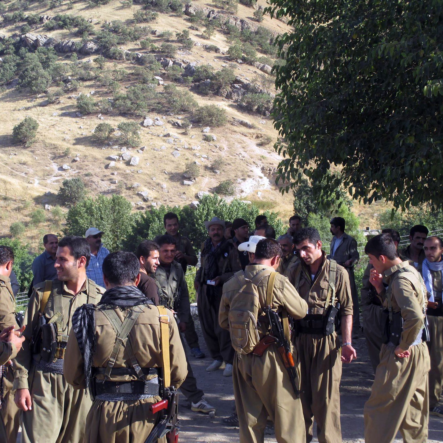 الصورة تظهر مجموعة من الرجال يرتدون ملابس عسكرية في منطقة طبيعية. توجد تلال خضراء في الخلفية، وبدت بعض الأشجار. يبدو أن هؤلاء الرجال يتفاعلون مع بعضهم البعض، حيث هناك من يتحدث ومن يتواجد في مجموعات صغيرة. يظهر في الصورة أيضا بعض الأشخاص يحملون أسلحة. الجو العام يوحي بنشاط اجتماعي أو تحضير لشيء ما.