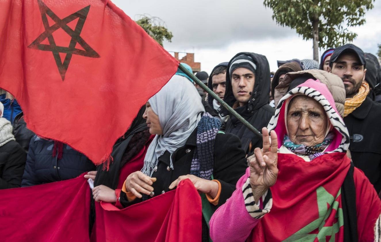 L'image montre une foule rassemblée, avec des gens portant des drapeaux marocains. Parmi eux, on peut voir une femme âgée qui fait un signe de paix avec sa main. Les participants semblent engagés et soutiennent une cause commune, probablement en lien avec leur identité ou des revendications sociales. Leur expression et leur posture suggèrent une forte détermination et un esprit de solidarité. Il y a une atmosphère de mobilisation collective.