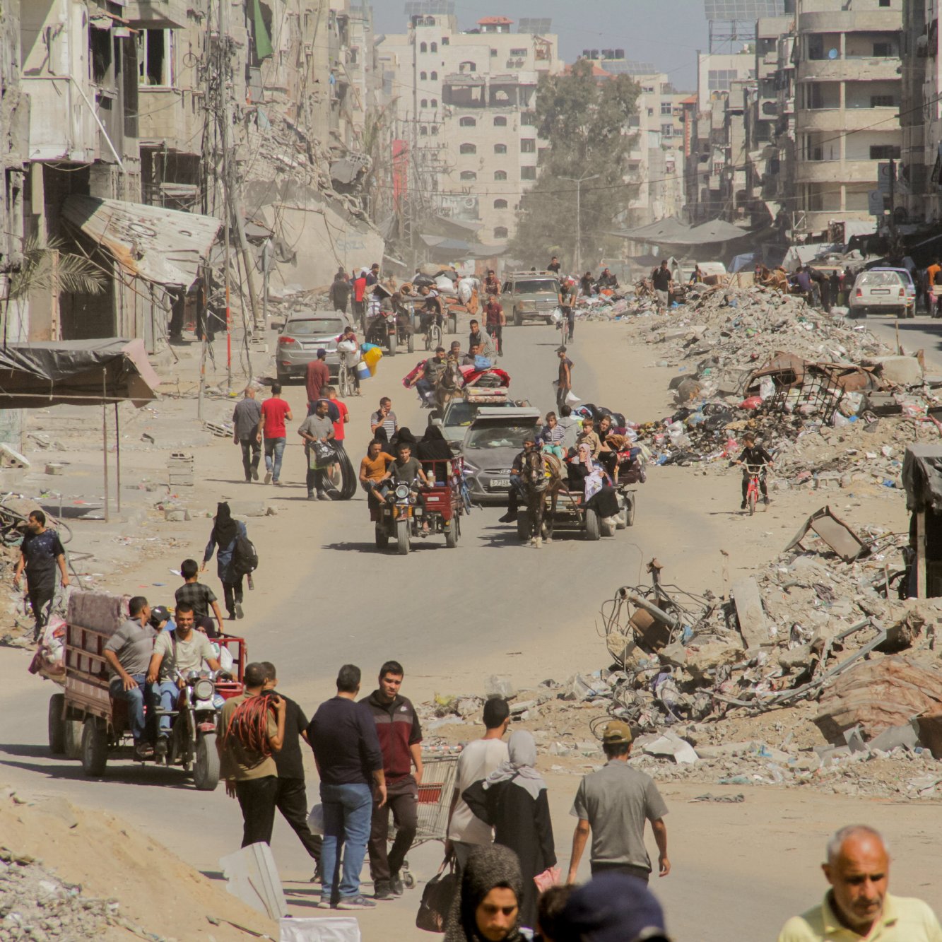 L'immagine mostra una scena di una città con edifici distrutti e rovine visibili. La strada è affollata da persone che camminano e da veicoli, inclusi motocicli e carretti. La polvere sollevata dall'ambiente circostante crea un'atmosfera di desolazione. In lontananza si vedono ulteriori segni di distruzione, con detriti sparsi lungo i lati della strada. L'insieme comunica un forte senso di difficoltà e resilienza in un contesto di crisi.