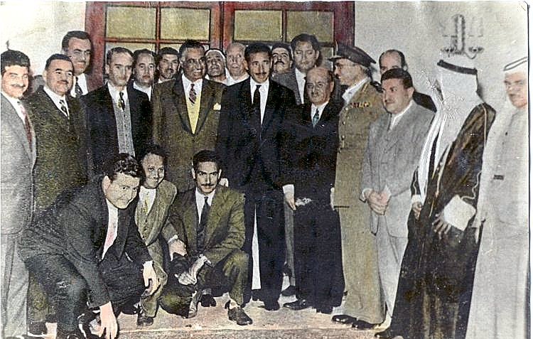 L'image montre un groupe de plusieurs hommes rassemblés dans une pièce. Ils portent des vêtements variés, allant de costumes formels à des tenues traditionnelles. Certains se tiennent debout tandis que d'autres sont accroupis ou assis. Le décor semble être d'une certaine importance, potentiellement une cérémonie ou un événement officiel. Les expressions faciales des personnes varient, allant de sourires à des regards sérieux, ce qui suggère un mélange de convivialité et de gravité. Les détails sur les personnalités présentes et le contexte de l'image ne sont pas précisés.