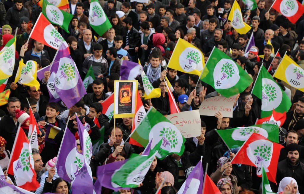 L'image montre une foule de manifestants brandissant des drapeaux colorés, principalement des couleurs violet, vert et rouge, qui semblent appartenir à un mouvement politique. Certaines personnes portent des affiches ou des pancartes. L'atmosphère semble dynamique et engagée, avec une concentration sur la participation collective. Les visages des manifestants sont variés et montrent un mélange de détermination et d'unité. Les drapeaux, avec des symboles, suggèrent une forte identité politique.