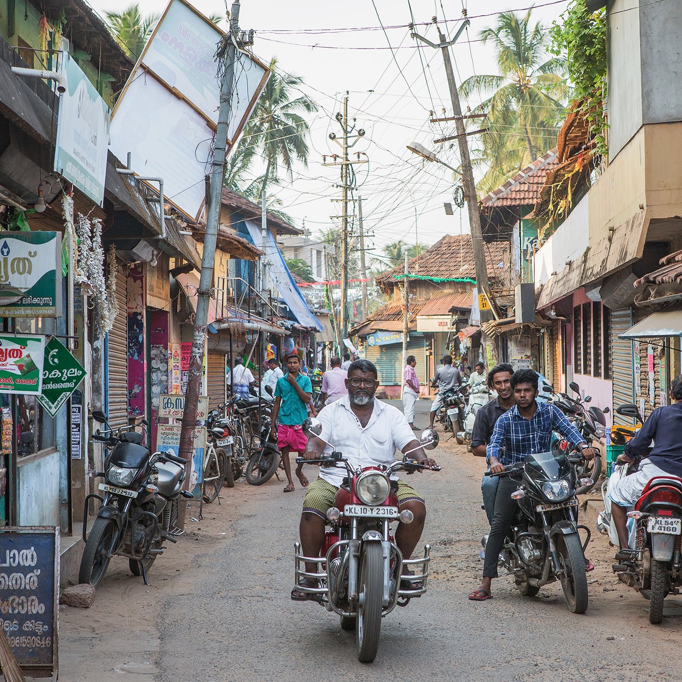 تظهر الصورة مشهداً من شارع ضيق في منطقة ريفية أو شبه حضرية. يتواجد في الصورة عدد من الأشخاص يسيرون ويتحدثون، بينما يقود رجل دراجة نارية في منتصف الشارع. المباني على الجانبين بسيطة ومكونة من طوابق قليلة، مع أسطح مغطاة بالبلاط. يمكن رؤية لافتات ملونة على المحلات التجارية التي تبيع مختلف السلع، وهناك أشجار تتواجد في الخلفية مما يعطي انطباعاً بحياة محلية نابضة. الجو يبدو هادئاً ومعتاداً على النشاط اليومي للسكان.