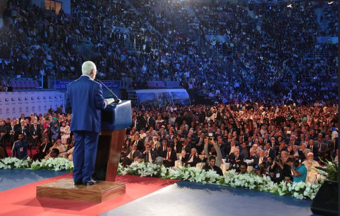 الصورة تظهر تجمعاً كبيراً في قاعة اجتماعات أو مؤتمر، حيث يقف متحدث أمام منصة ويبدو أنه يلقي خطابًا. الحضور بكثافة يجلسون في مقاعدهم، وتحيط بهم أضواء جميلة. يوجد تزيين بالأزهار البيضاء على الجانبين، مما يضفي جواً احتفائياً. الجو العام يوحي بأهمية الحدث واهتمام كبير من الجمهور.