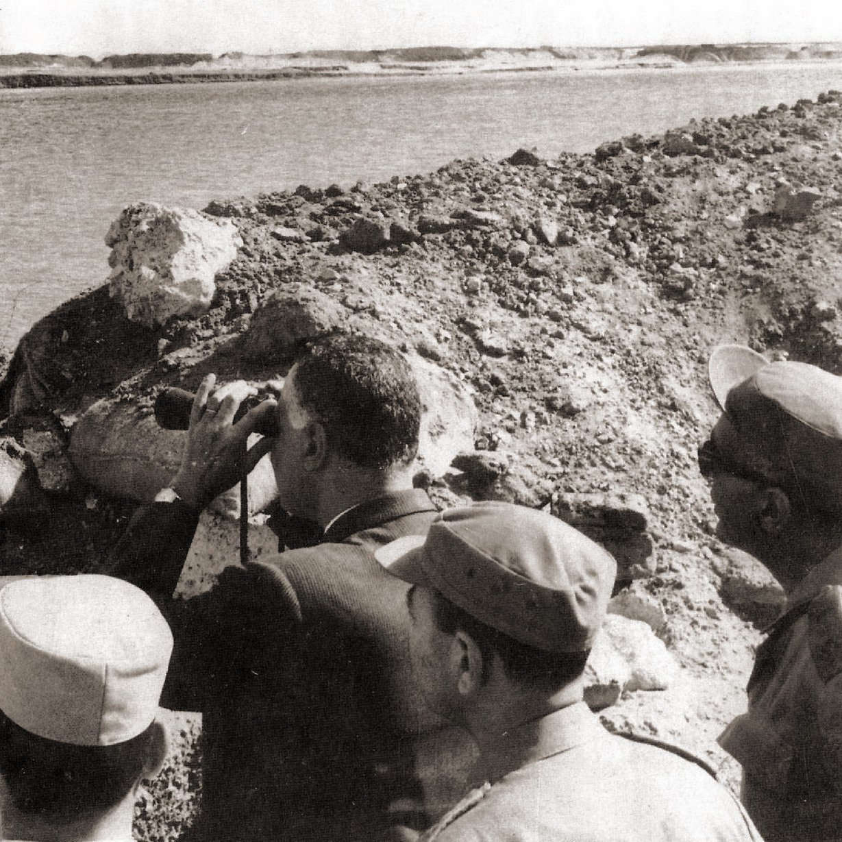 L'image montre un groupe d'hommes observant un paysage, probablement au bord d'une rivière ou d'un lac. L'un des hommes, qui semble en costume, utilise des jumelles pour scruter l'horizon. Les autres sont en tenue militaire et semblent attentifs à ce qui se passe devant eux. L'environnement est aride, avec des collines de terre et peu de végétation visible en arrière-plan. Cette scène évoque une situation de surveillance ou d'évaluation d'un site stratégique.