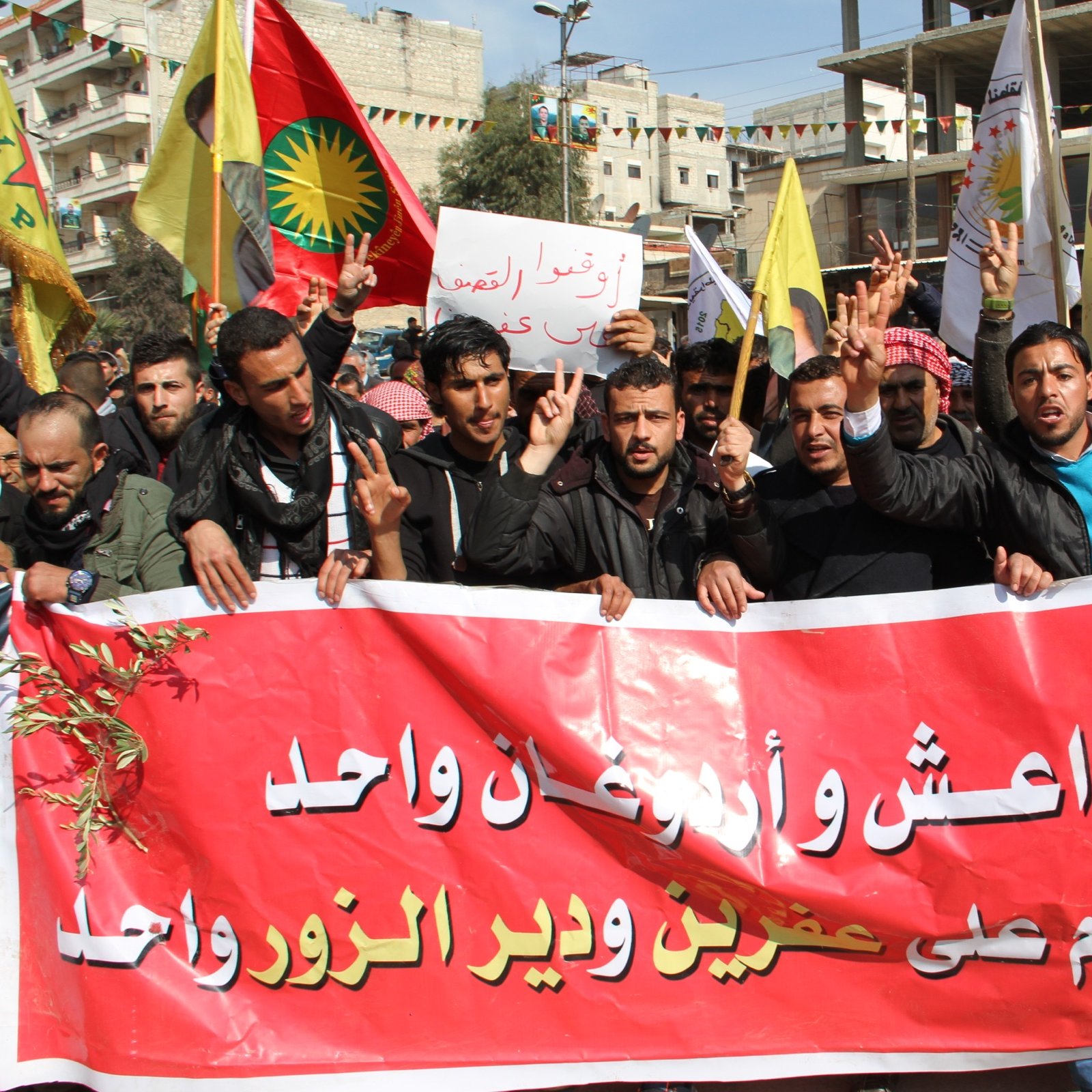 L'image montre un groupe de manifestants tenant des pancartes et des drapeaux, en train de défiler dans une rue. Ils portent des expressions déterminées et semblent militer pour une cause. Leurs vêtements et les drapeaux indiquent une provenance régionale. Une grande banderole rouge est affichée, avec des inscriptions en arabe. Les manifestants font le signe de la victoire avec leurs doigts, ce qui suggère un esprit de solidarité et de lutte. L'ambiance générale semble dynamique et engagée.