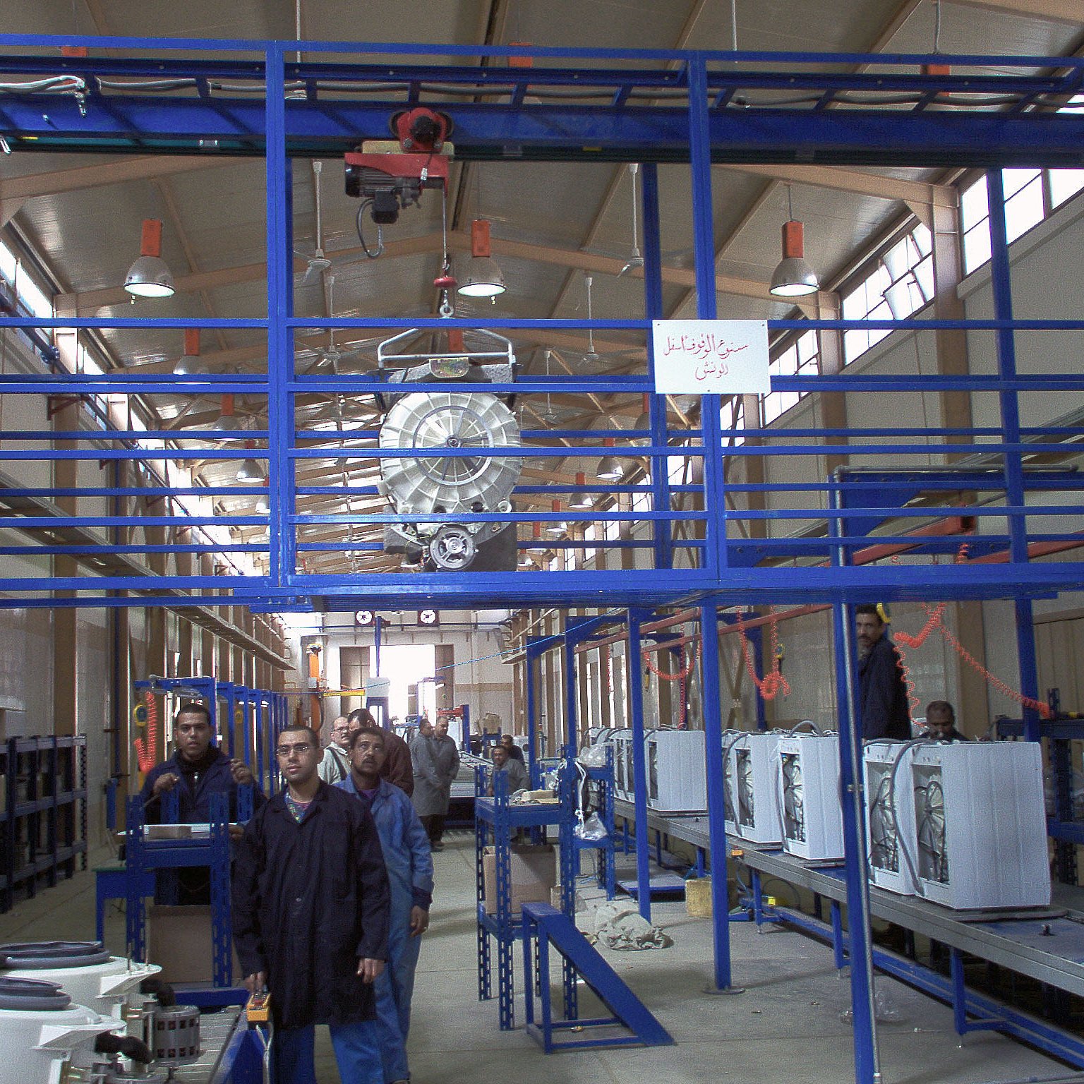 L'image montre un environnement d'atelier ou d'usine. On peut y voir des structures en métal, probablement des systèmes de rangement ou de support. Il y a des personnes travaillant dans cet espace, accompagnées d'équipements comme des appareils électroménagers, qui semblent être en phase de montage ou de préparation. L'éclairage est suffisant, et l'agencement des machines et des travailleurs suggère une ligne de production ou un processus d'assemblage. On note également des éléments rigides de la construction qui maintiennent l'espace organisé.