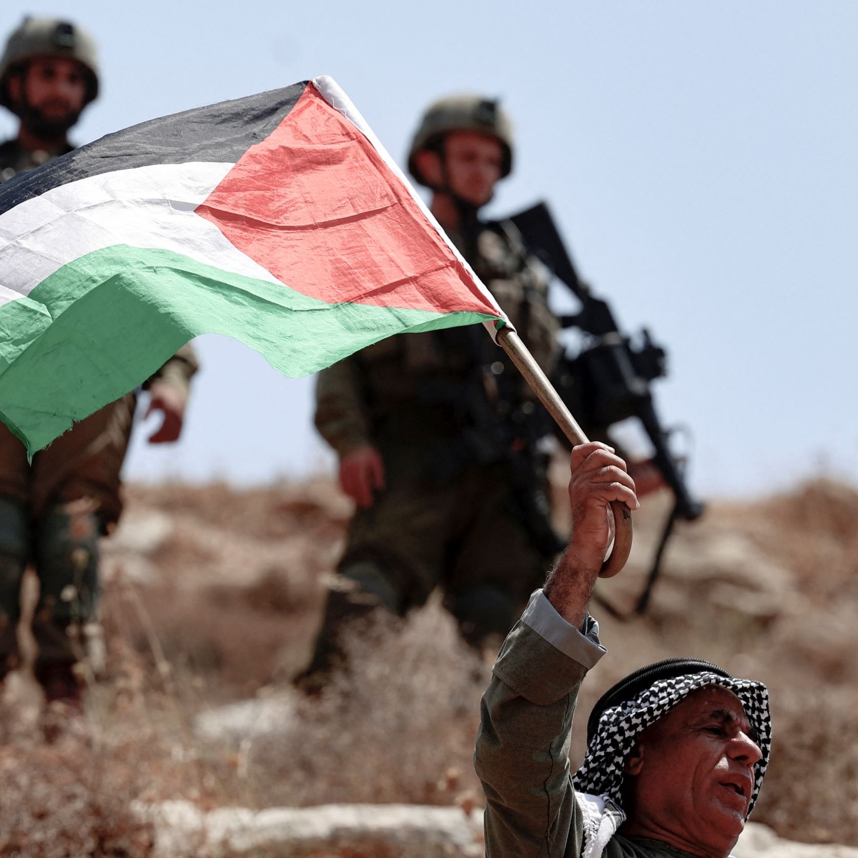 The image shows a man holding a Palestinian flag aloft, standing in a rocky landscape. He appears to be expressing a strong patriotic sentiment. In the background, a group of soldiers is visible, dressed in military gear, suggesting a tense or conflict-related scene. The setting looks arid, typically associated with certain regions in Palestine. The overall atmosphere conveys a sense of tension and resistance.