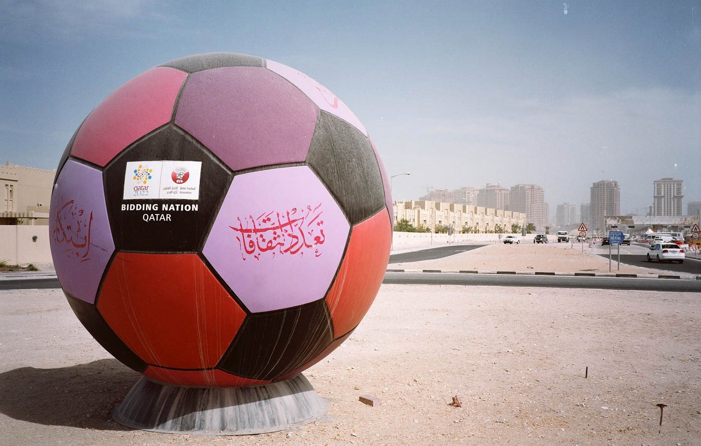 L'image montre un énorme ballon de football décoré, affichant des inscriptions colorées en arabe et d'autres éléments graphiques. Ce ballon est installé sur un terrain, probablement dans un contexte urbain, avec des bâtiments en arrière-plan. Le ciel est clair, et l'environnement semble être une zone en développement ou un lieu préparé pour un événement, suggérant une connexion avec le football ou un événement sportif marquant, tel qu'une compétition internationale.