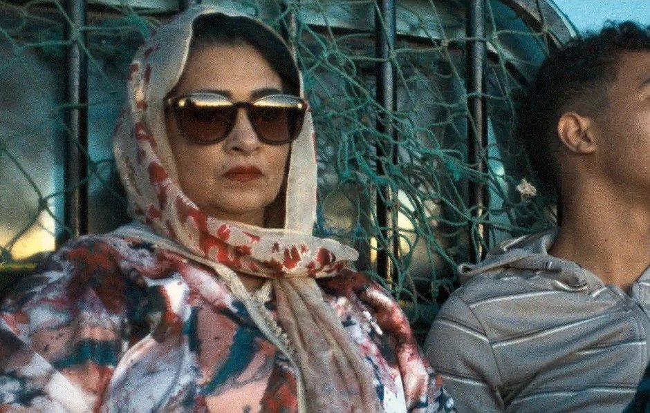 L'image montre une femme et un jeune homme assis ensemble dans un environnement désertique. La femme porte un foulard et des lunettes de soleil, affichant une expression attentive. Elle est vêtue d'une veste aux motifs colorés. À côté d'elle, le jeune homme porte un sweat à capuche et semble pensif, regardant au loin. En arrière-plan, on aperçoit des éléments qui évoquent un paysage aride. L'éclairage donne une atmosphère calme et contemplative à la scène.