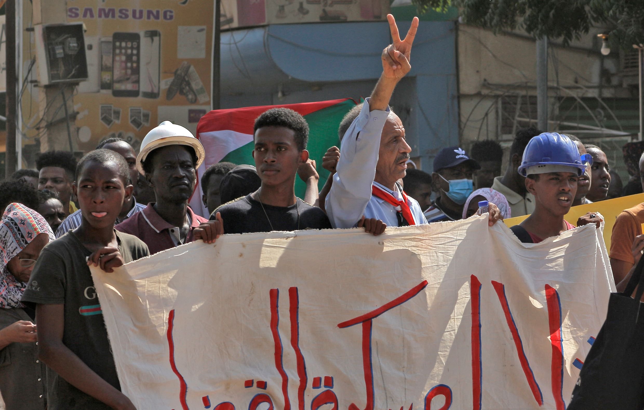 در این تصویر، گروهی از معترضان در حال برگزاری یک تظاهرات هستند. یکی از افراد، در حالی که علامت پیروزی را بالا برده، توجه را به خود جلب کرده است. دیگر شرکت‌کنندگان نیز با چهره‌های جدی و مصمم در کنار او ایستاده‌اند. در دست آنها یک بنر بزرگ دیده می‌شود که به زبان عربی نوشته شده و احتمالاً به اعتراضات مرتبط است. پس‌زمینه تصویر شامل ساختمان‌ها و تابلوهای تبلیغاتی است. احساس کلی تصویر نشان‌دهنده روحیه فعالان اجتماعی و خواسته‌های آنهاست.