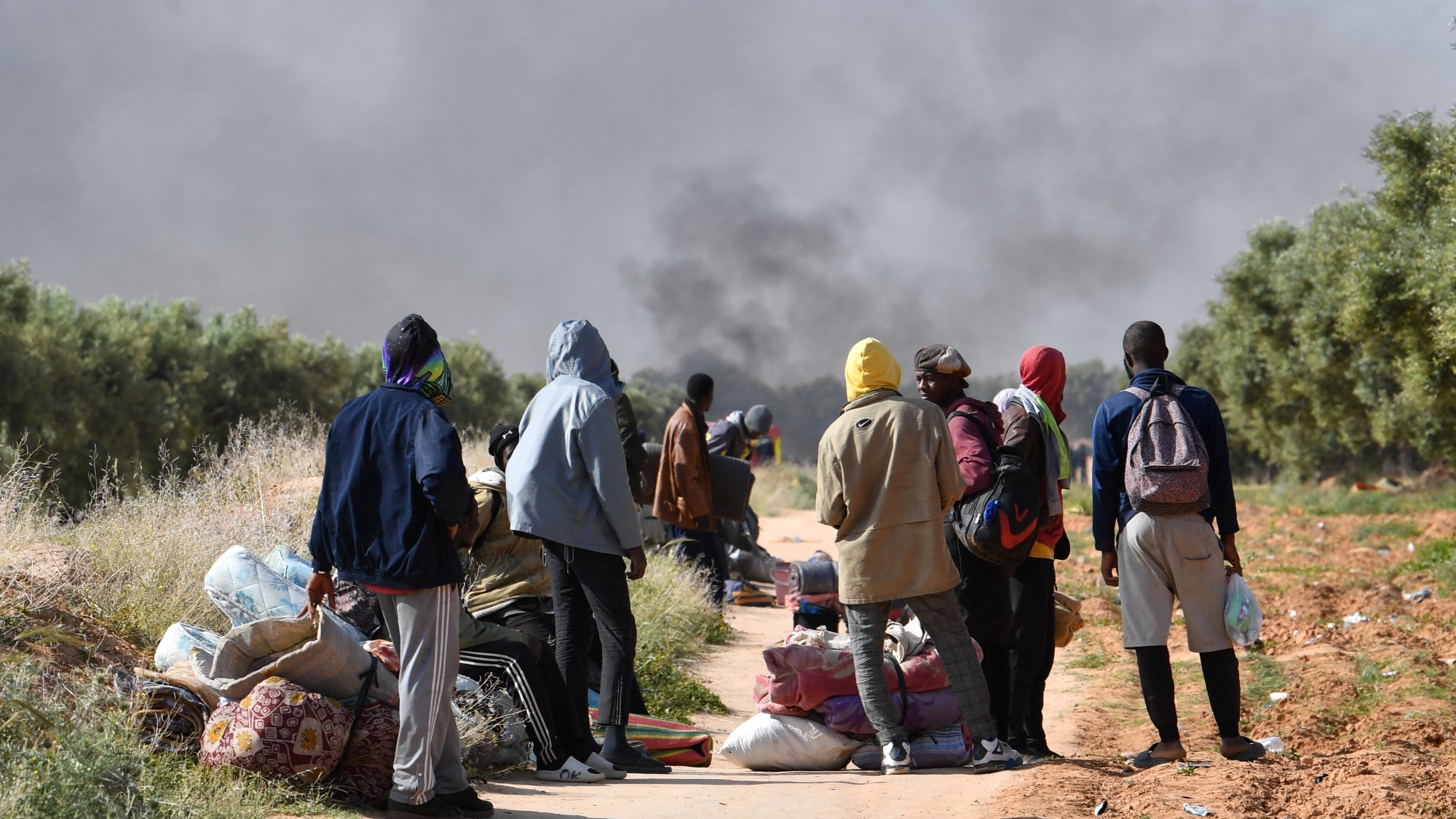 Un groupe de personnes se tient sur un chemin, entouré de fumée et de bagages.