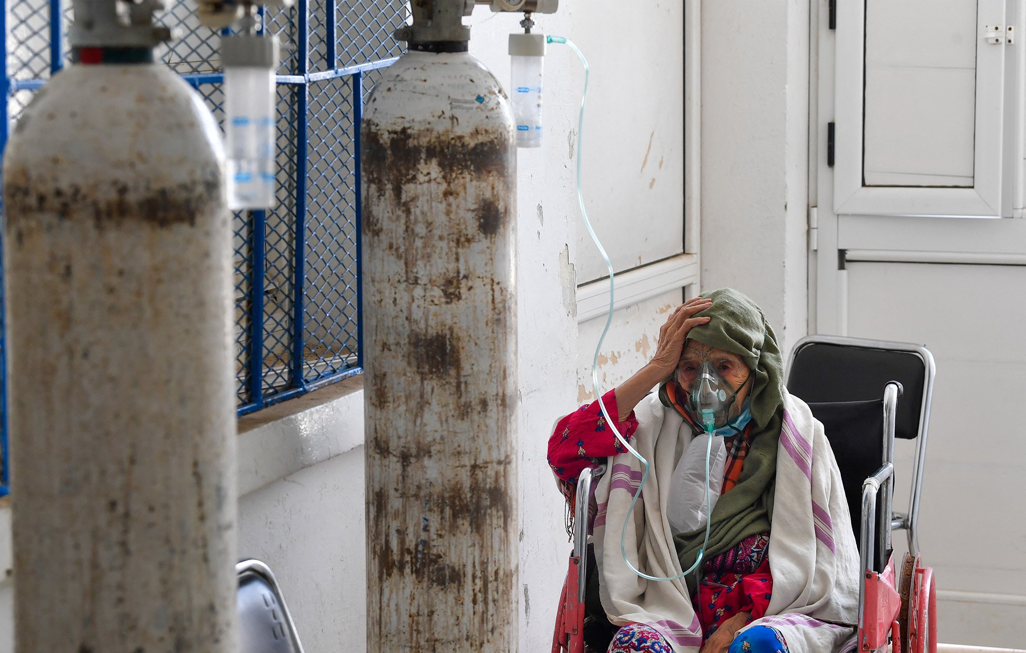 L'immagine mostra una donna anziana seduta su una sedia a rotelle in un ambiente ospedaliero. Indossa una maschera per l'ossigeno e sembra preoccupata, con una mano sulla fronte. Dietro di lei ci sono bombole di ossigeno e un ambiente che appare abbastanza semplice e clinico. L'atmosfera trasmette una sensazione di vulnerabilità e dolore, evidenziando le difficoltà legate alla salute.