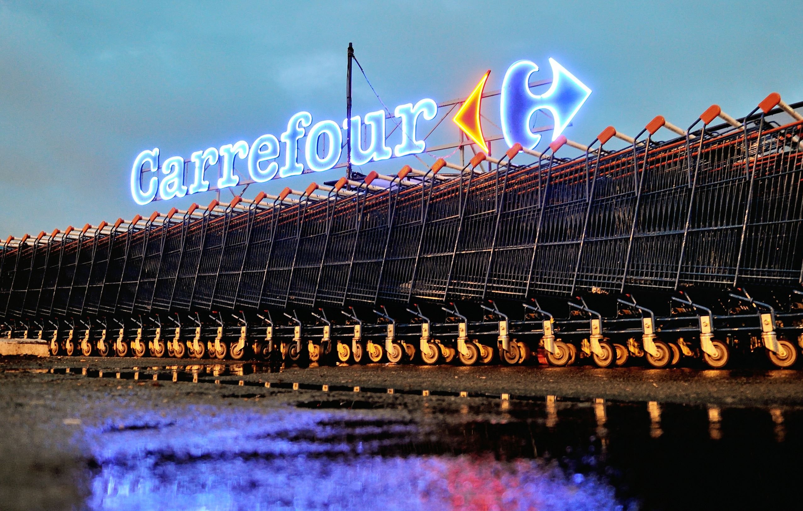 L'image montre un rayonnement lumineux du logo de Carrefour, un hypermarché bien connu, en arrière-plan. Devant le logo, il y a une série de caddies alignés sur le sol, probablement après la pluie, ce qui crée des reflets sur la surface. L'atmosphère est calme et nocturne, accentuée par l'éclairage des enseignes. Les caddies sont organisés de manière ordonnée, offrant une vue intéressante sur l'environnement d'un magasin de grande distribution.