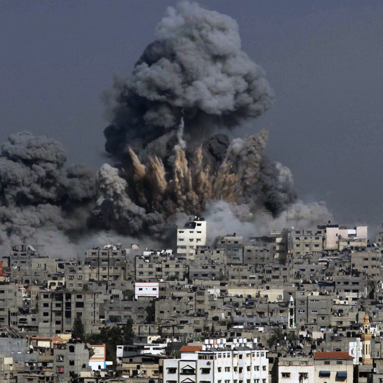 الصورة تظهر مشهدًا لمدينة تعرضت لتفجير، حيث تندلع سحب كثيفة من الدخان الأسود في السماء. يظهر تأثير الانفجار في المباني المحيطة، حيث تبرز آثار الدمار، بينما يبدو أن السماء ملبدة بالغيوم الناتجة عن الانفجار. المدينة تبدو كثيفة البنايات مع تشوهات واضحة نتيجة الأحداث المدمرة.