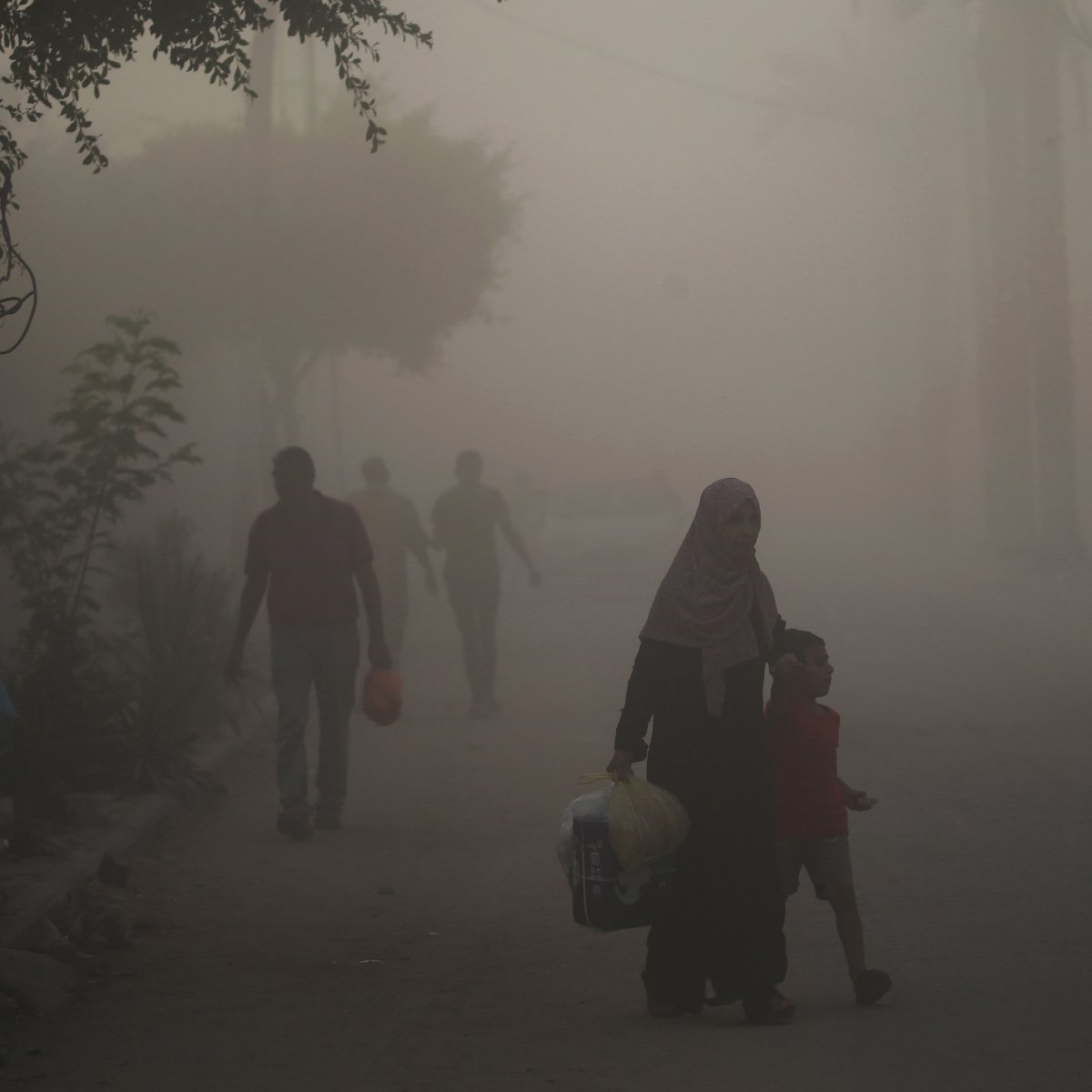 L'image montre une scène urbaine plongée dans une atmosphère brumeuse ou poussiéreuse. Dans le premier plan, une femme portant un hijab tient la main d'un jeune garçon. Ils semblent marcher ensemble le long de la rue. En arrière-plan, d'autres silhouettes de personnes sont visibles, floues par la brume, tandis que des arbres et des structures urbaines apparaissent à peine à travers la poussière. L'ambiance semble calme mais chargée d'un certain mystère à cause de la visibilité réduite.