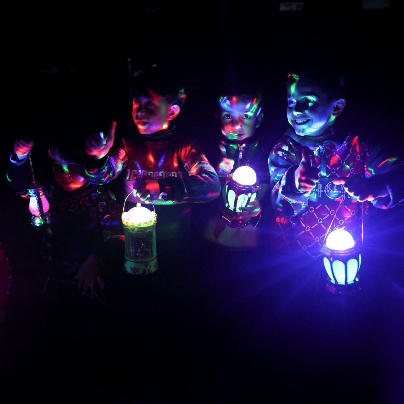 L'image montre un groupe d'enfants dans l'obscurité, tenant des lanternes colorées qui émettent des lumières vives. Les lumières créent des reflets et des motifs multicolores sur leurs visages et leurs vêtements. Les enfants semblent joyeux et amusés, s'amusant avec les lanternes pendant la nuit. L'ambiance est festive et magique grâce aux jeux de lumière.