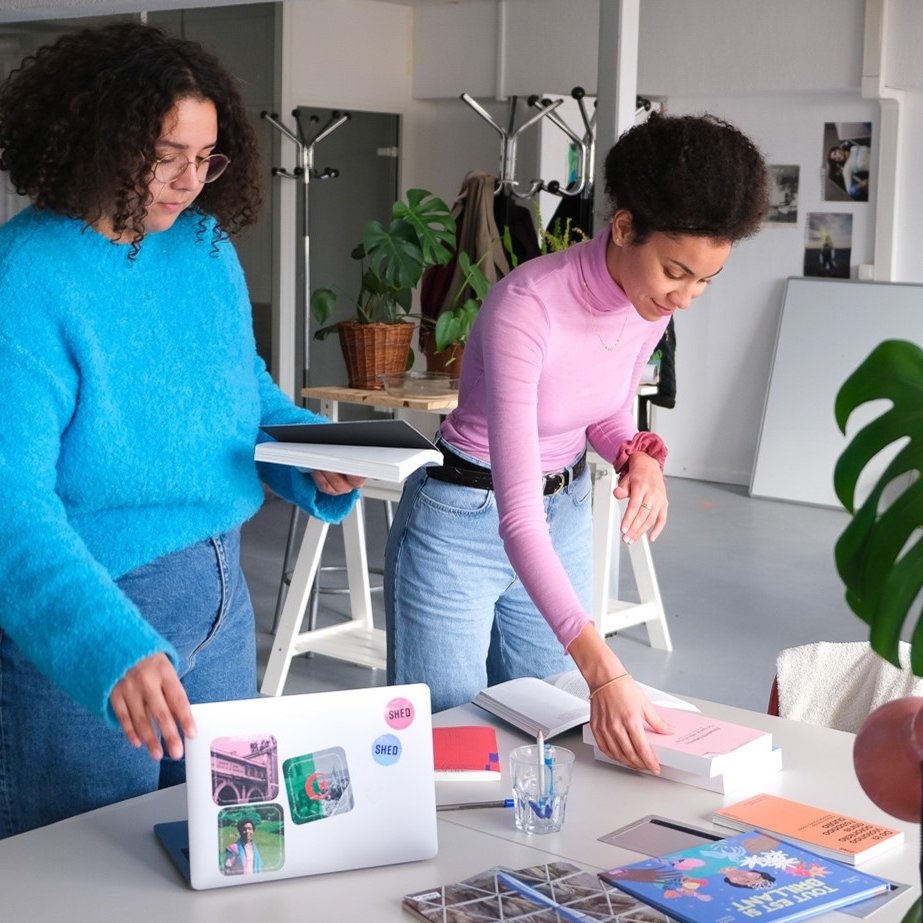 En la imagen se pueden ver a dos mujeres en un espacio de trabajo moderno y luminoso. Una de ellas, con cabello rizado y vestido azul, está sosteniendo unos cuadernos o carpetas, mientras que la otra, con cabello rizado y recogido, está organizando unos libros sobre una mesa. En la mesa hay una computadora portátil, algunos materiales impresos y una planta decorativa. El ambiente parece creativo y acogedor, con elementos de diseño contemporáneo y luminosidad natural.