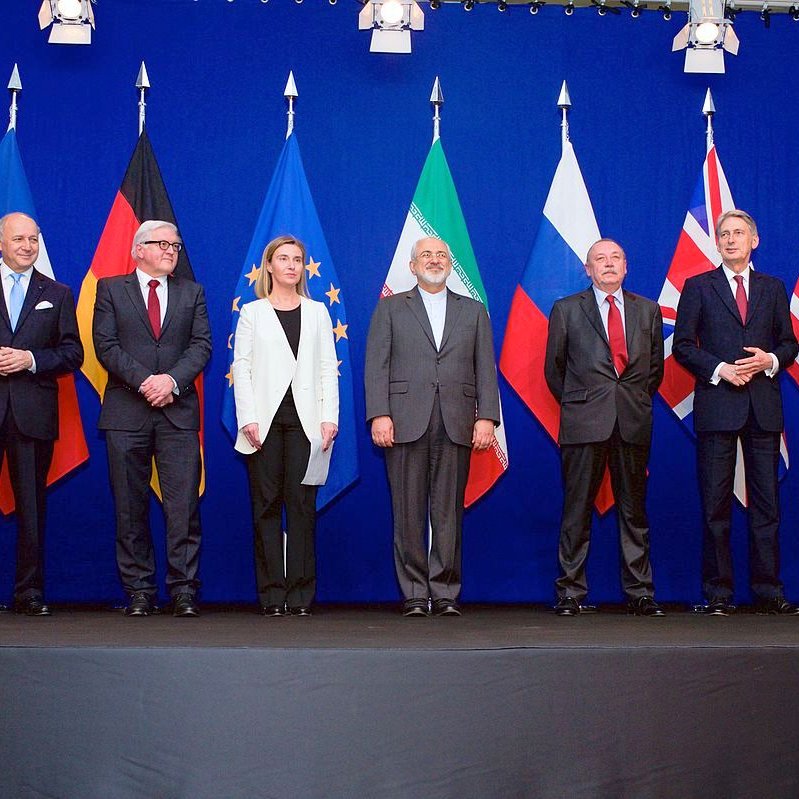 L'image montre un groupe de personnalités politiques posant ensemble devant un mur de drapeaux. On peut voir des représentants de plusieurs pays avec des drapeaux de la Chine, de l'Allemagne, de l'Union européenne, de l'Iran, de la Russie, du Royaume-Uni et des États-Unis en arrière-plan. Les personnes semblent discuter d'un sujet important et portent des costumes formels. L'ambiance est sérieuse, correspondant à un événement diplomatique.