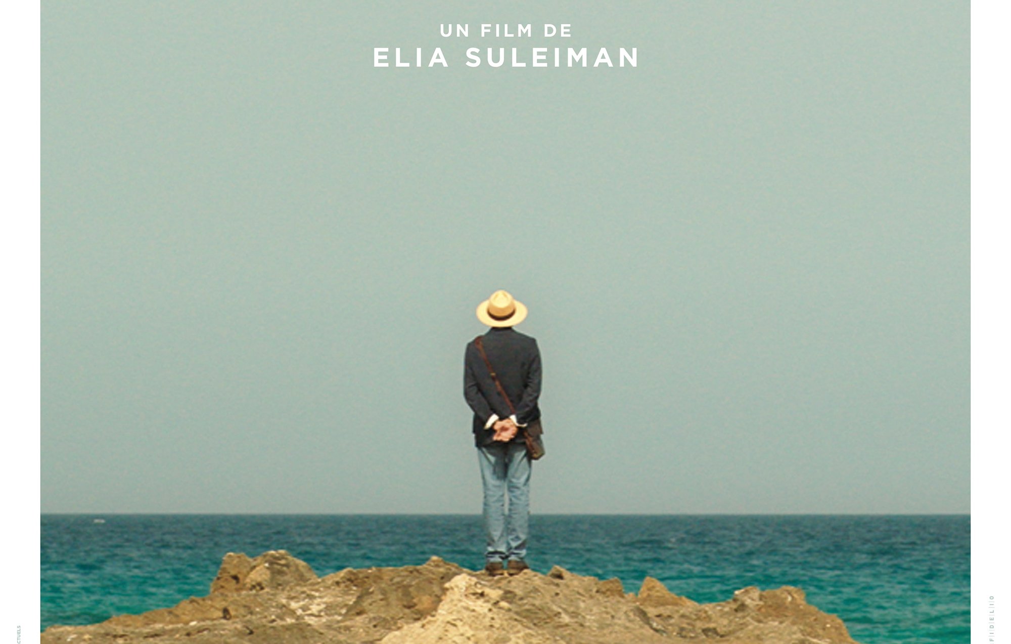 L'image représente une affiche de film intitulée "It Must Be Heaven", réalisé par Elia Suleiman. On y voit un homme de dos, portant un chapeau, se tenant debout sur un rocher au bord de la mer. L'horizon est dégagé, avec un ciel bleu clair. Le titre du film est inscrit en lettres blanches en bas de l'affiche, avec également le nom du réalisateur. L'ensemble de l'image évoque une ambiance contemplative et poétique.