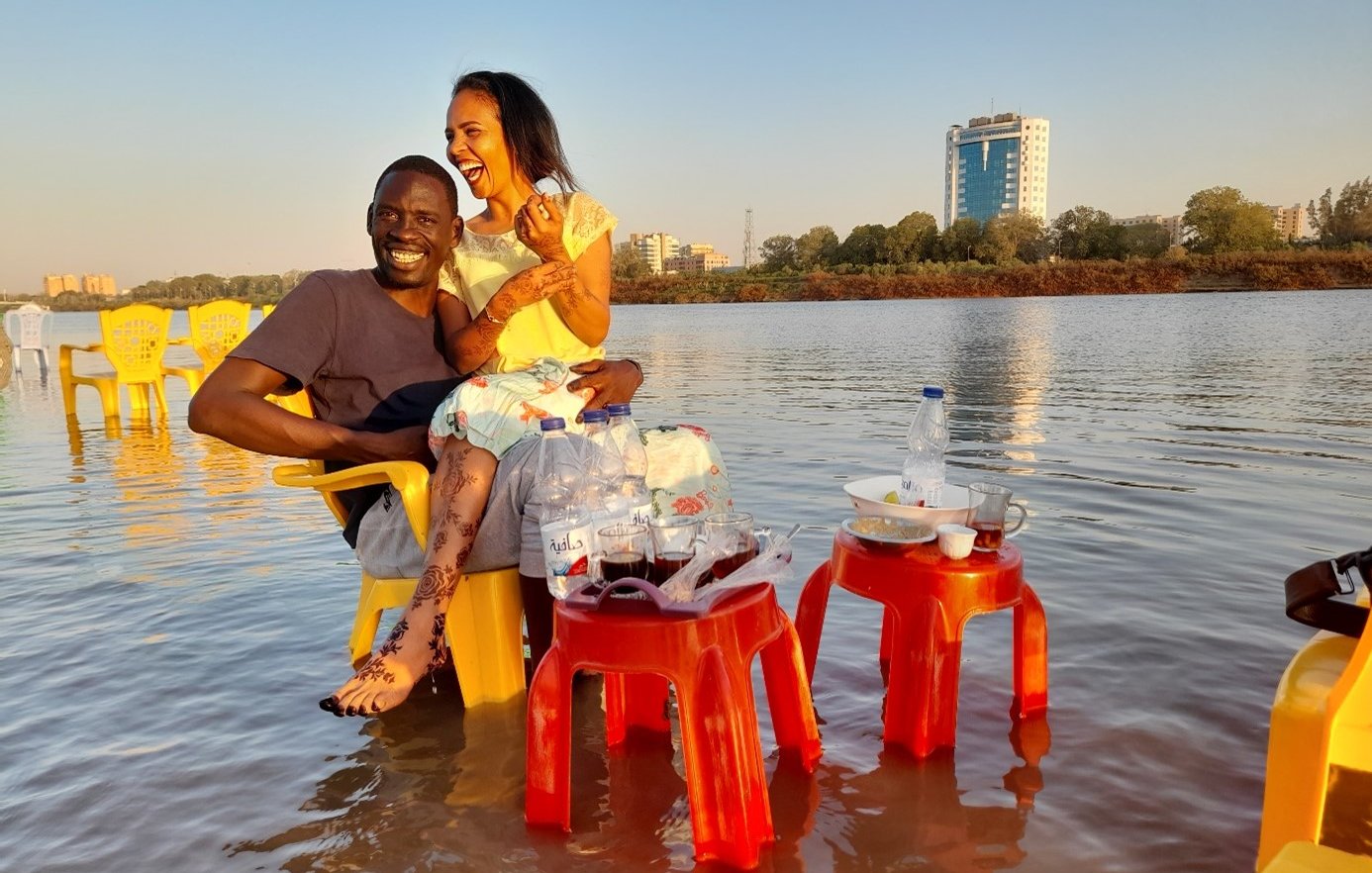 L'image montre un couple assis sur des chaises jaunes au bord de l'eau. Ils semblent heureux et détendus, avec un grand bâtiment en arrière-plan. Des bouteilles d'eau et des tasses sont posées sur des petites tables à proximité. Le cadre est serein, avec un ciel dégagé et l'eau qui reflète la lumière du soleil, créant une ambiance chaleureuse.