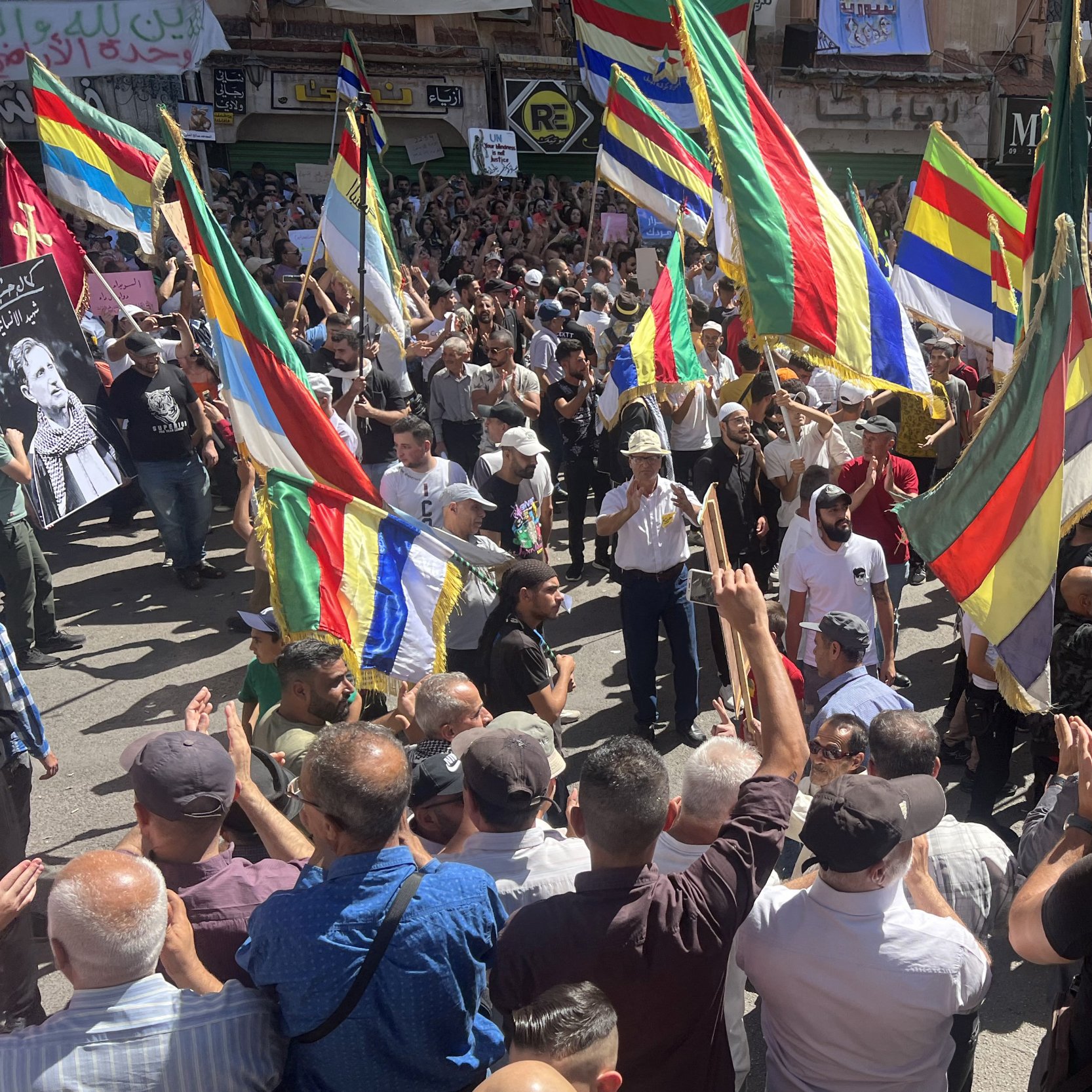L'image montre une grande foule rassemblée lors d'une manifestation. Les participants tiennent des drapeaux colorés, probablement de différentes communautés ou groupes. On peut voir des visages de personnes connues affichés sur des pancartes. L'atmosphère semble dynamique et engagée, avec des manifestants applaudissant et exprimant leurs opinions. Le décor urbain suggère que l'événement se déroule dans une ville animée.