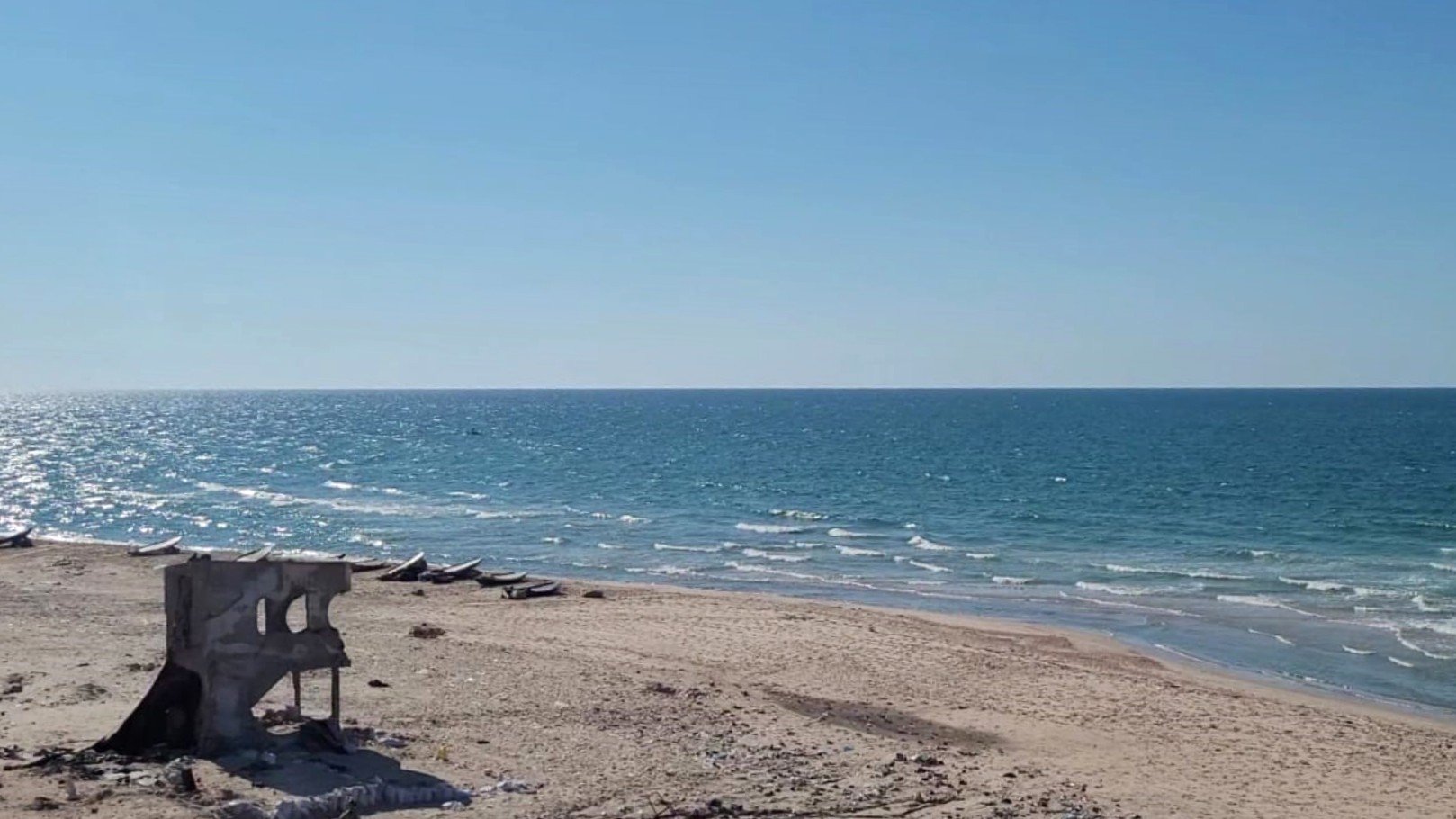 L'immagine mostra una spiaggia con sabbia chiara e un mare blu intenso sotto un cielo sereno e azzurro. In lontananza si possono vedere alcune imbarcazioni sulla superficie dell'acqua. Una struttura diroccata, grigia e in parte crollata, si trova sulla spiaggia, creando un contrasto con il paesaggio naturale circostante.