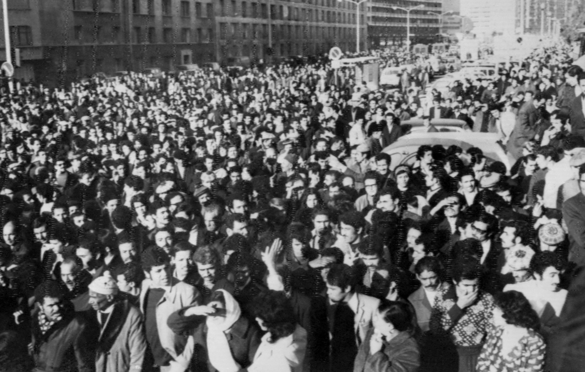 L'image montre une grande foule rassemblée dans une rue. Les personnes semblent se tenir très proches les unes des autres, créant une masse compacte. On peut apercevoir des bâtiments en arrière-plan, ainsi que des véhicules, ce qui indique que cette scène se déroule dans un milieu urbain. L'atmosphère semble intense et collective, suggérant un rassemblement pour une cause ou un événement significatif. Les vêtements des personnes, sombres et variés, donnent une impression de l'époque.