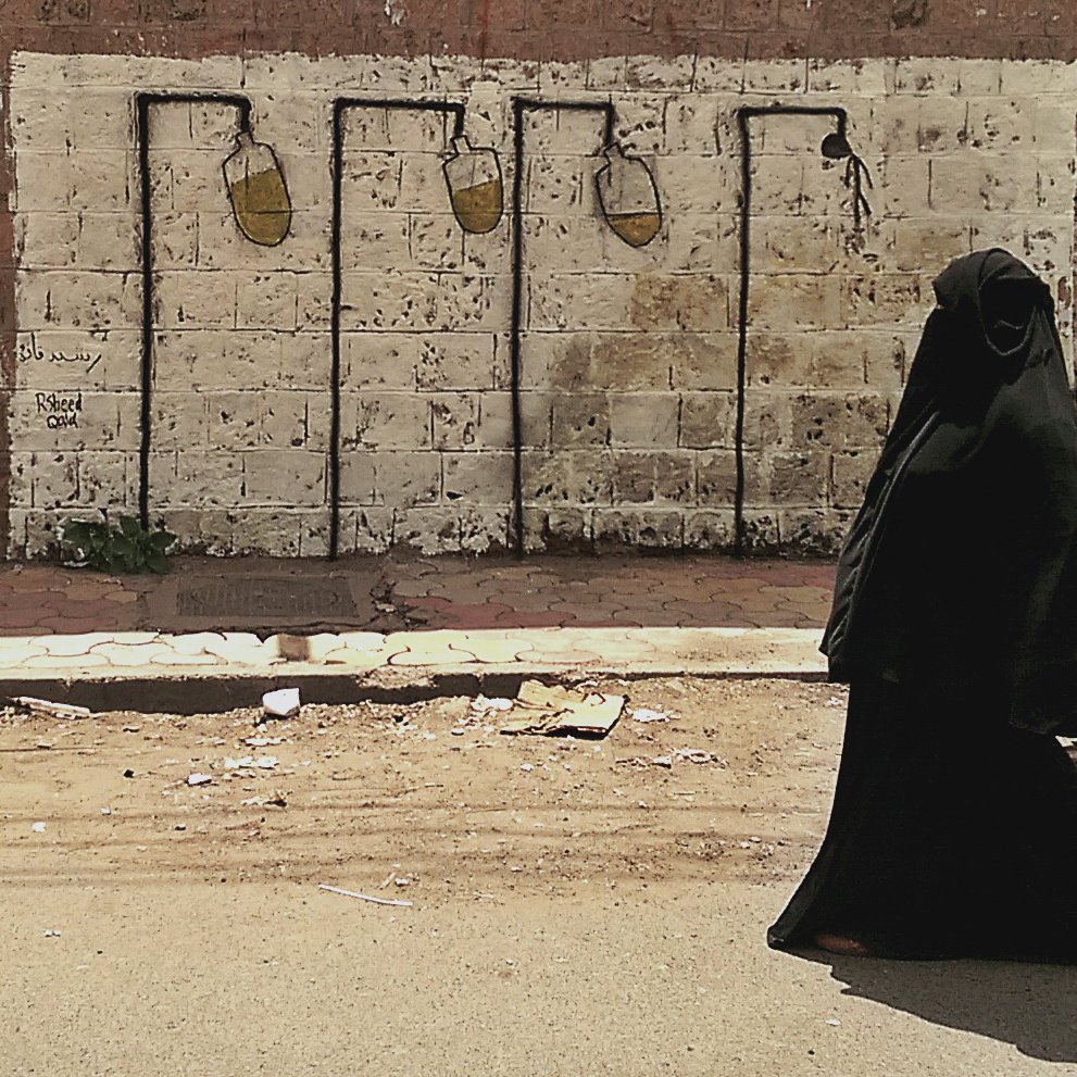 L'image montre une femme vêtue d'une tenue noire, probablement un hijab ou une burqa, marchant dans une rue. À ses côtés, un enfant tient sa main. En arrière-plan, on aperçoit un mur de béton avec un graffiti qui représente des pots de peinture suspendus, ajoutant une touche artistique à la scène. La route semble modeste avec des débris au sol, ce qui donne un aperçu de l'environnement urbain.