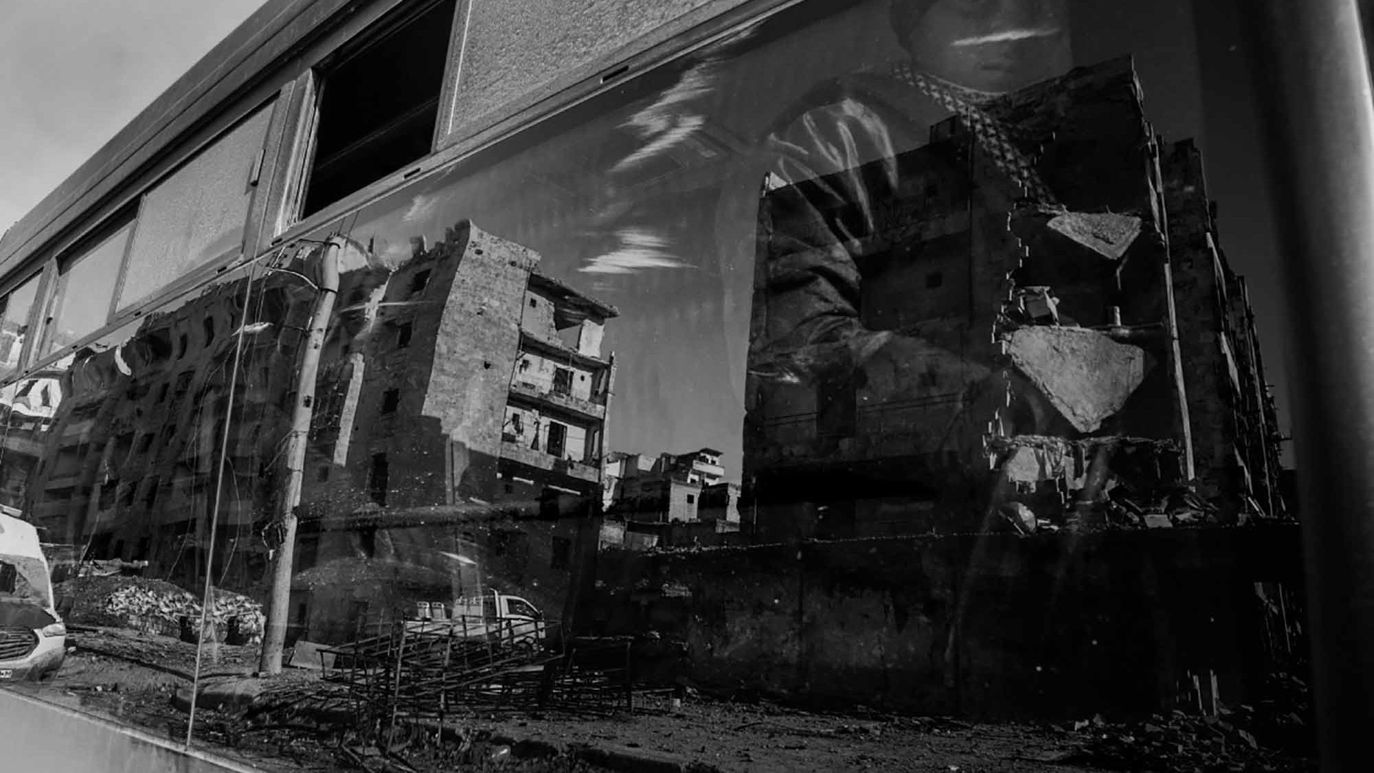 The image features a reflection captured on a glass surface, likely a window or a bus. It shows the distorted but vivid outlines of modern buildings that appear damaged or abandoned, suggesting a scene of urban decay. The reflection also includes a figure partially visible, wearing what looks like a patterned scarf or headdress. The overall tone of the image is monochromatic, emphasizing the somber, evocative nature of the scene. The contrast highlights the interplay between the busy life outside and the desolation depicted in the reflection.