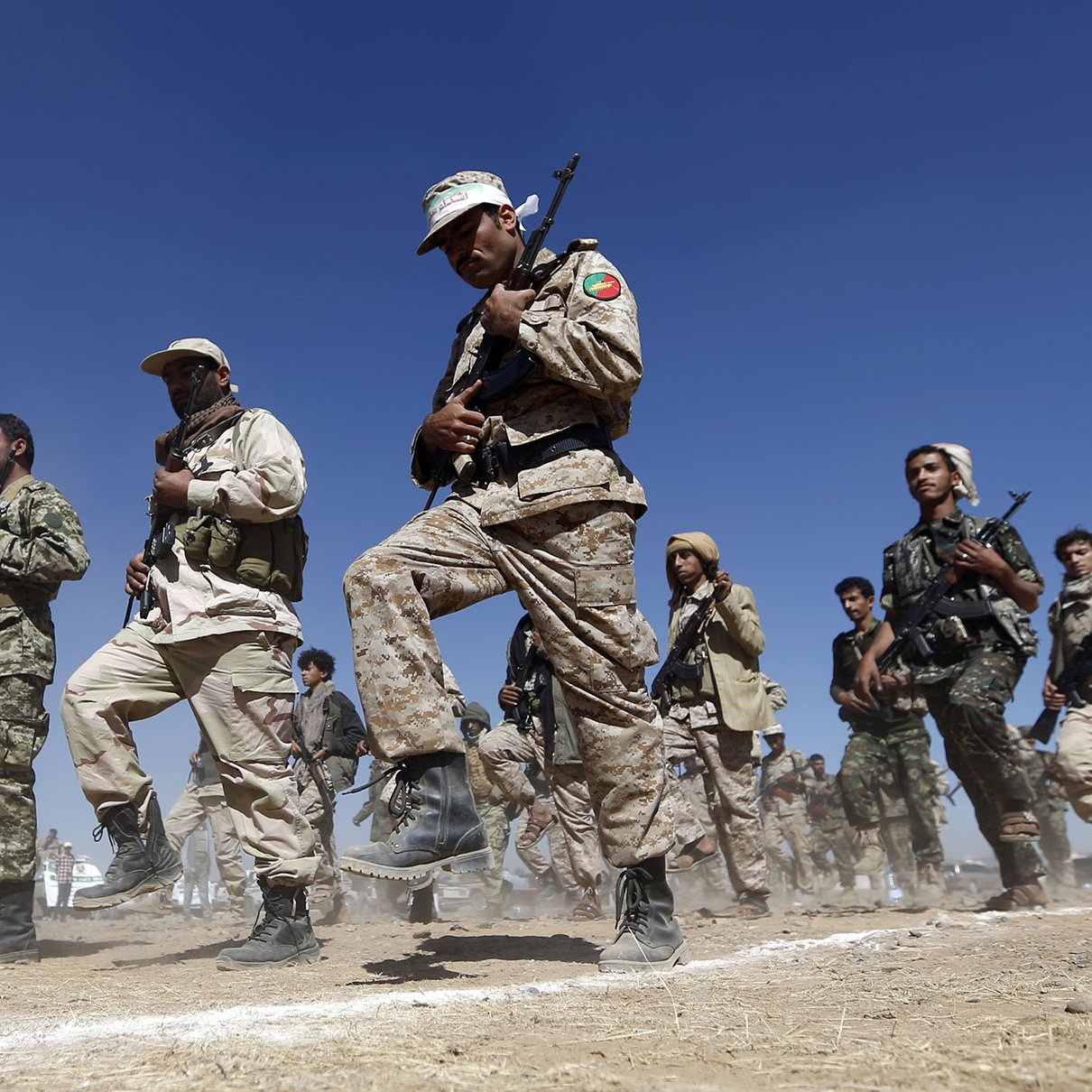 L'image montre un groupe de personnes en uniforme militaire en train de marcher ou de s'entraîner. Ils semblent être réunis en formation, tenant des fusils. Le terrain est poussiéreux, et on peut apercevoir un ciel bleu en arrière-plan. L'ambiance semble sérieuse et déterminée, suggérant une activité militaire ou un exercice de préparation.